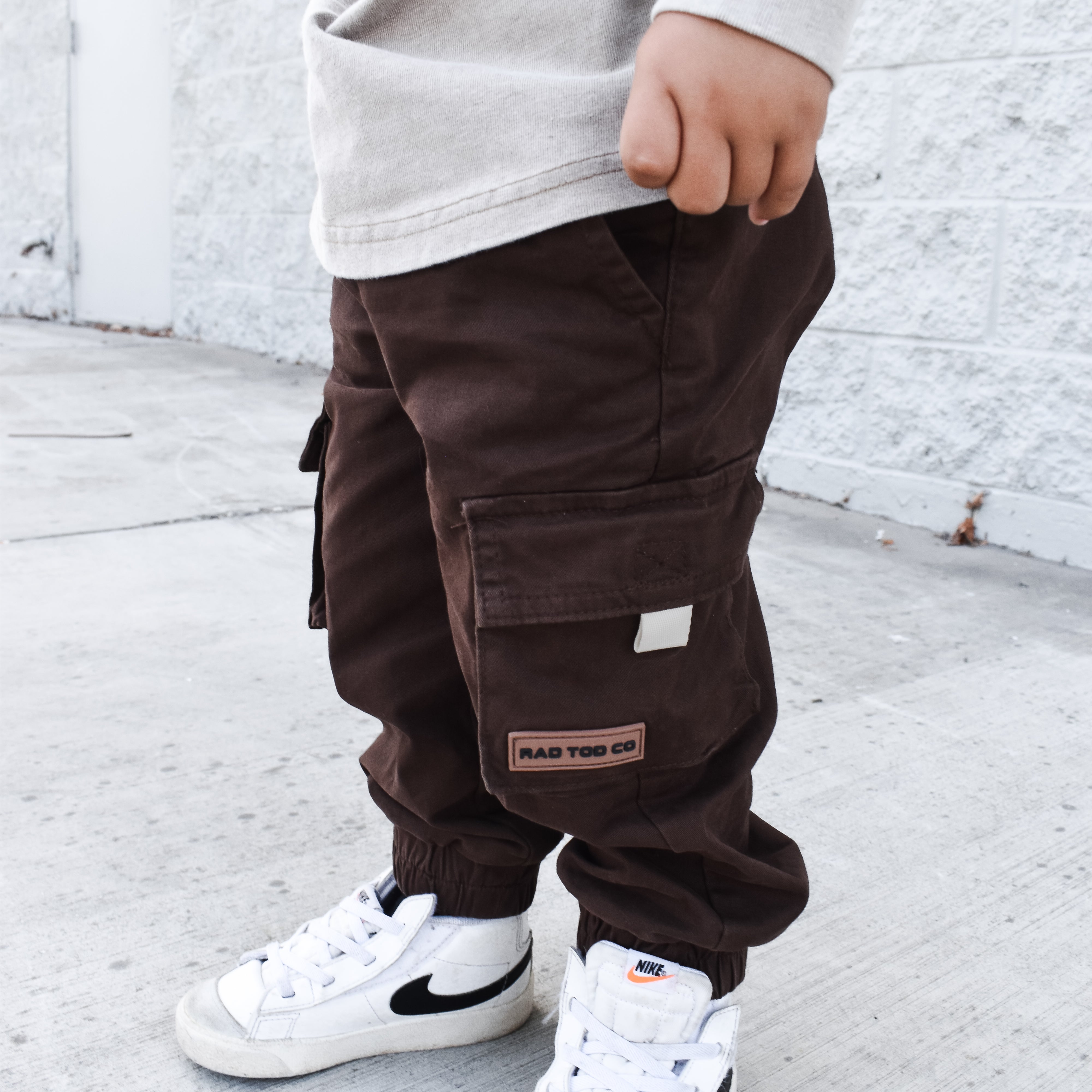 Child wearing brown cargo pants and white sneakers against a white brick wall.