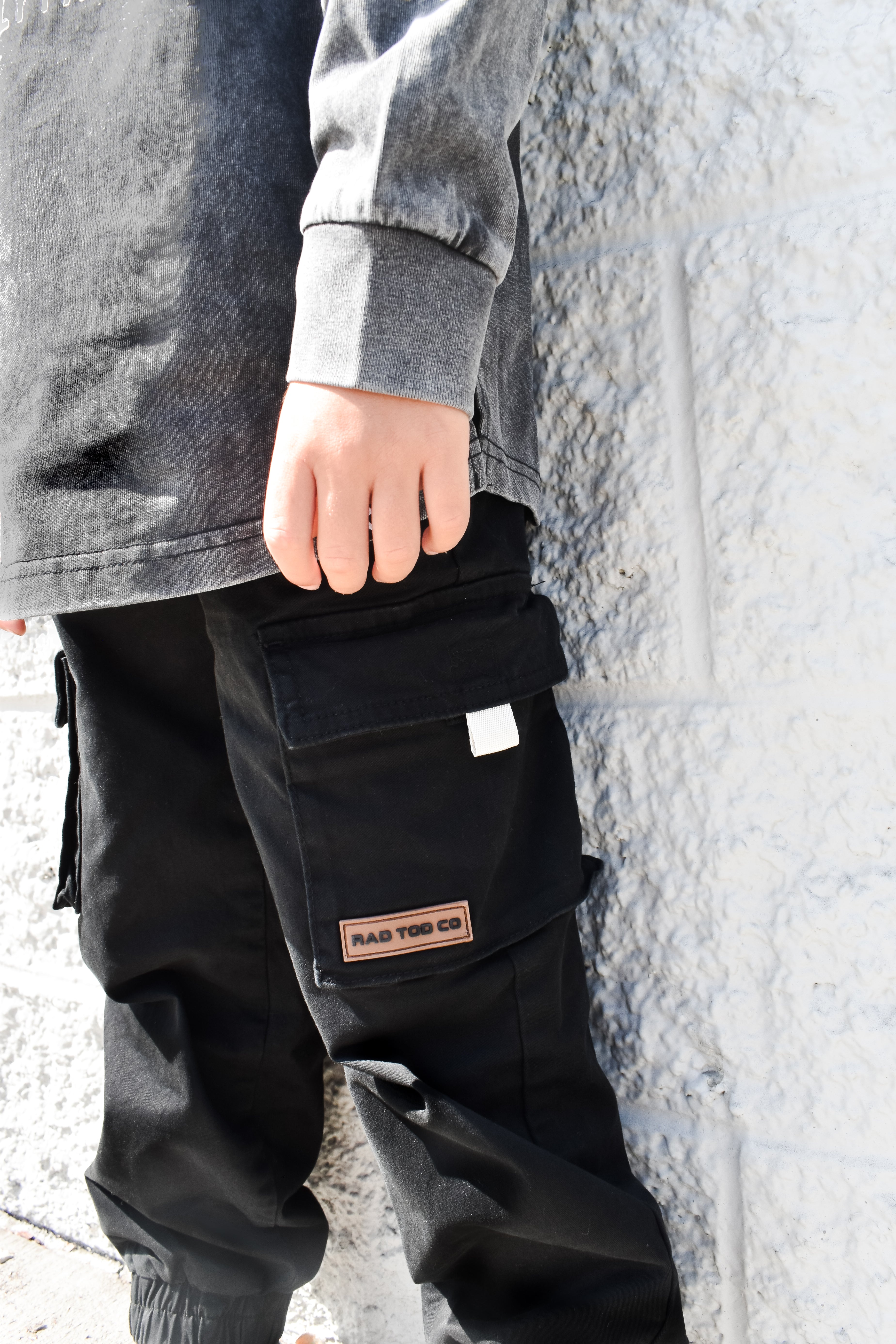 Person wearing black cargo pants by RAD TODDLER against a stone wall.