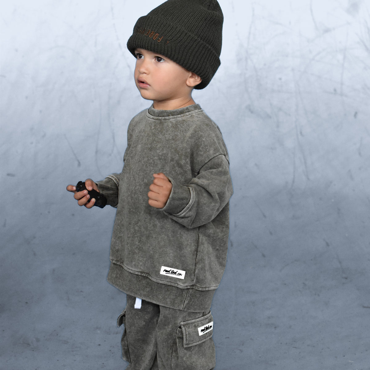 A toddler standing in profile wearing a dark olive green knit beanie, a matching washed olive green sweatshirt and cargo pants, and white sneakers against a textured gray studio backdrop.