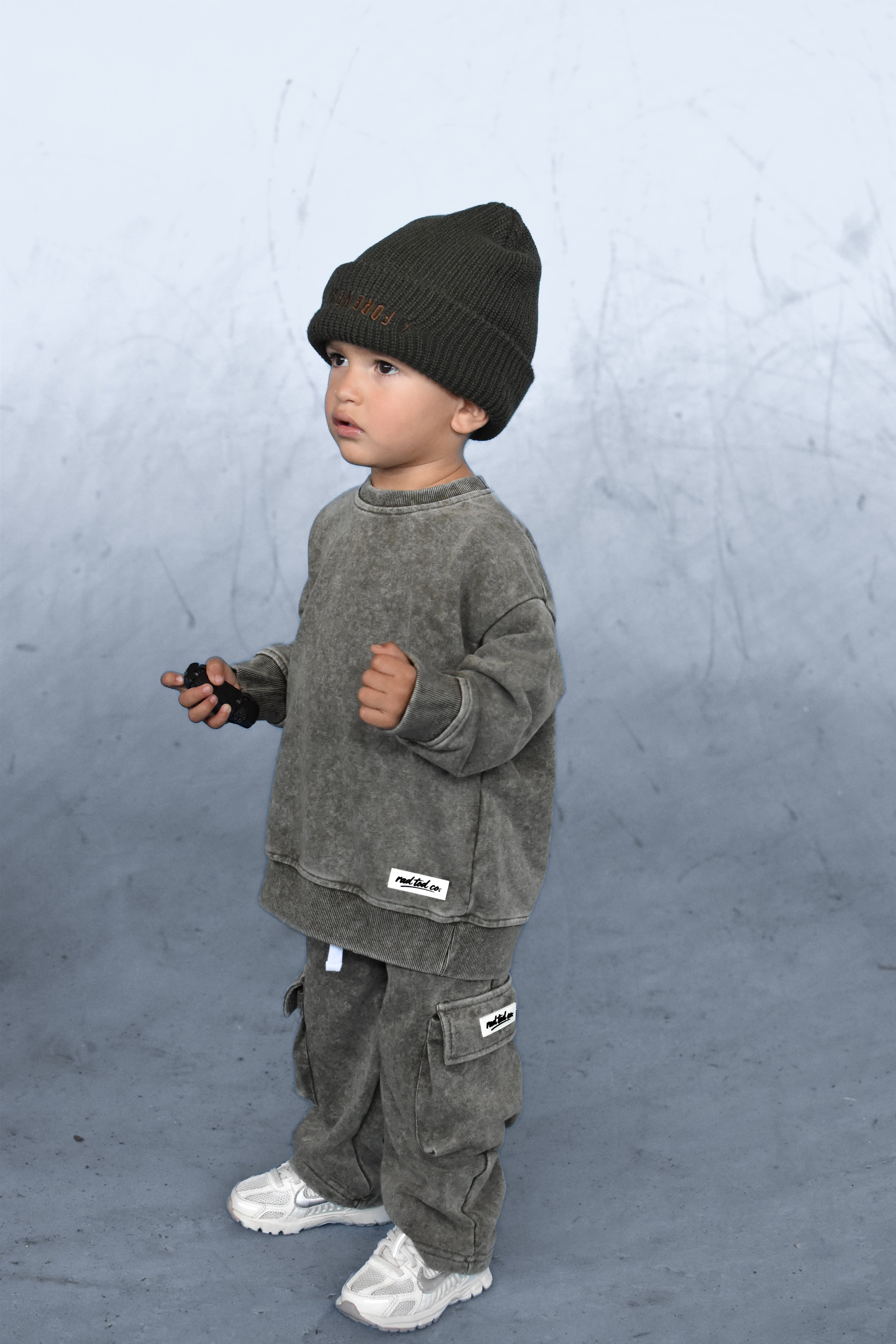 A toddler standing in profile wearing a dark olive green knit beanie, a matching washed olive green sweatshirt and cargo pants, and white sneakers against a textured gray studio backdrop.