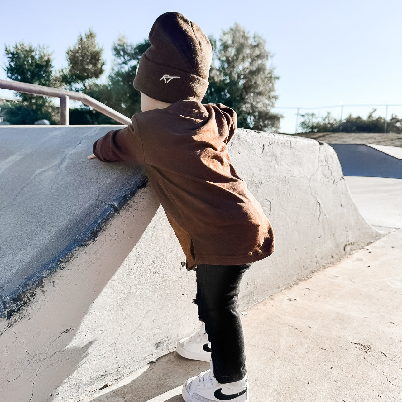 Child in a brown tee and black pants standing on a concrete ledge with trees in the background.