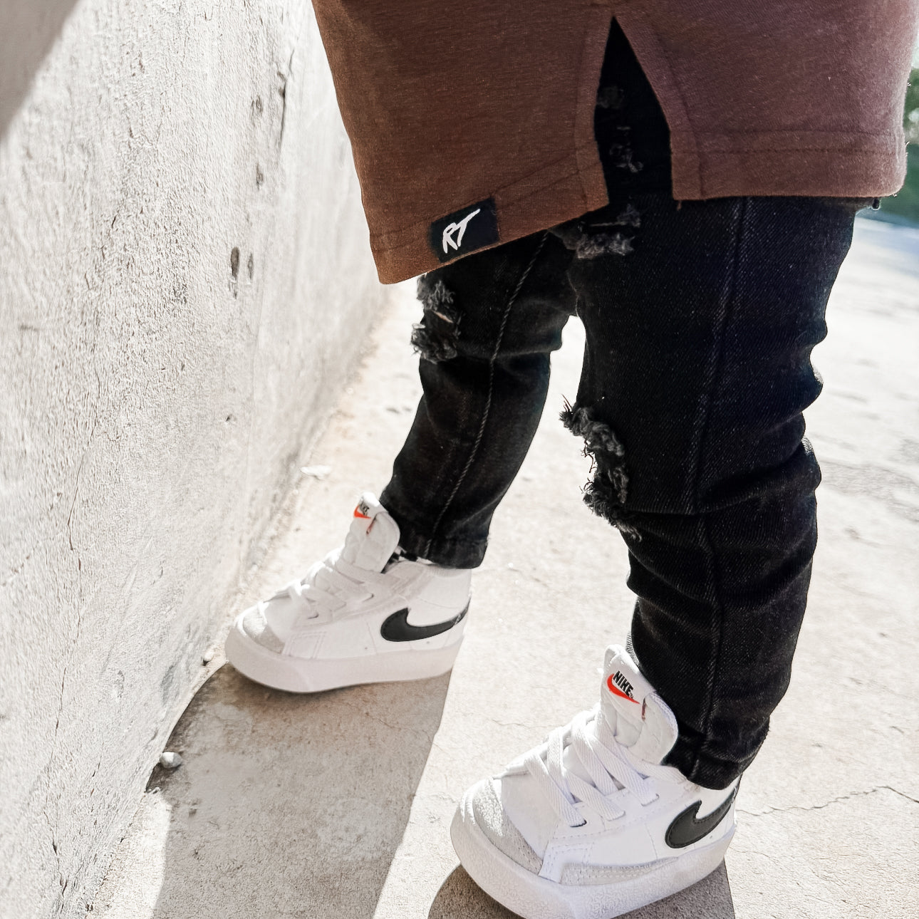 boy wearing a brown long sleeve tee, black ripped jeans, and white sneakers with a logo on a concrete surface.