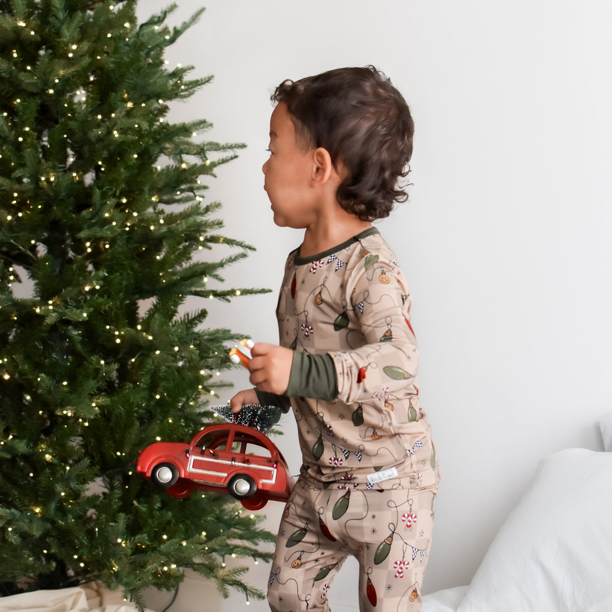 A toddler stands on a bed wearing a beige holiday-themed pajama set with a retro light print, holding a red toy car next to a decorated Christmas tree.