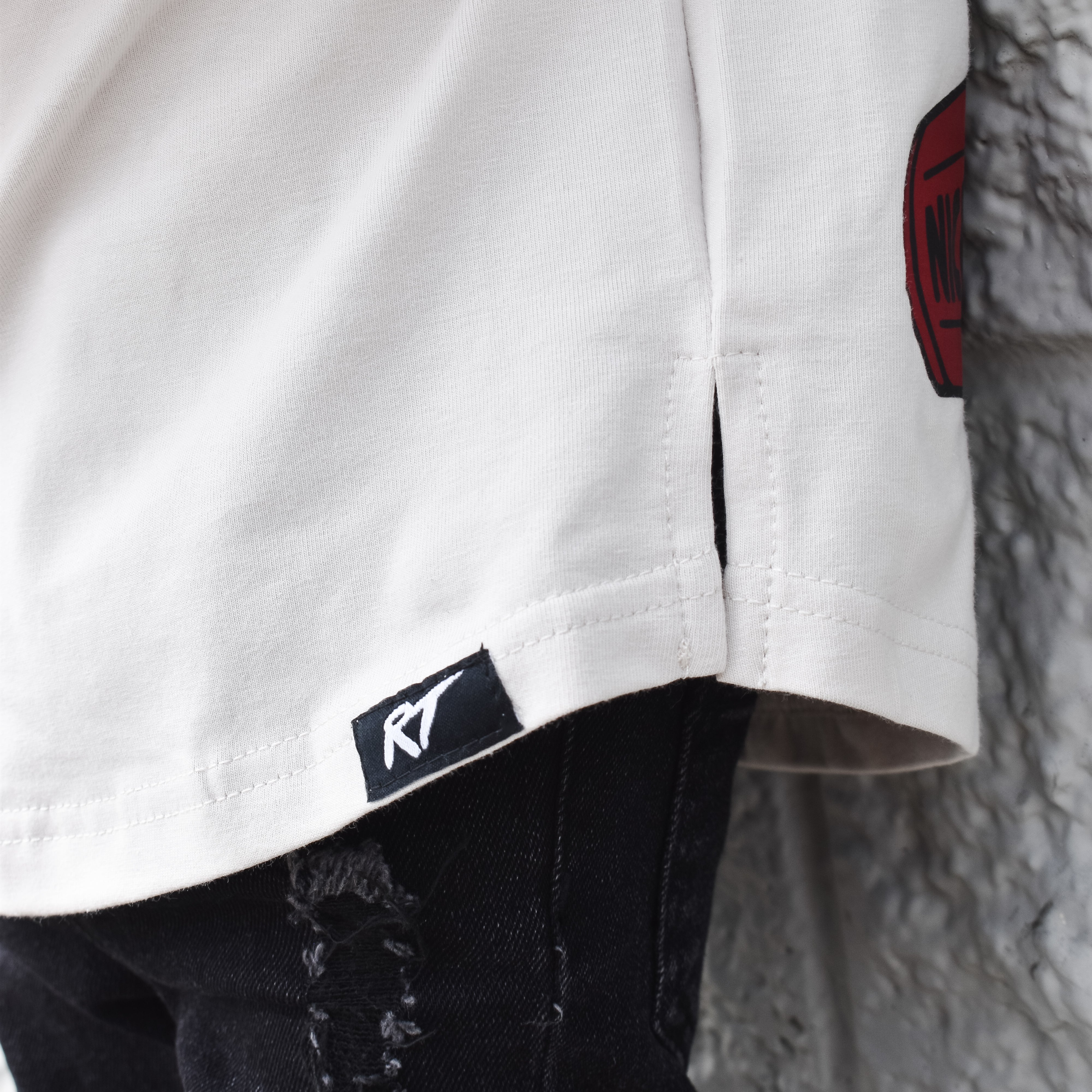 Close-up of a white shirt with a red logo and black pants against a textured wall.