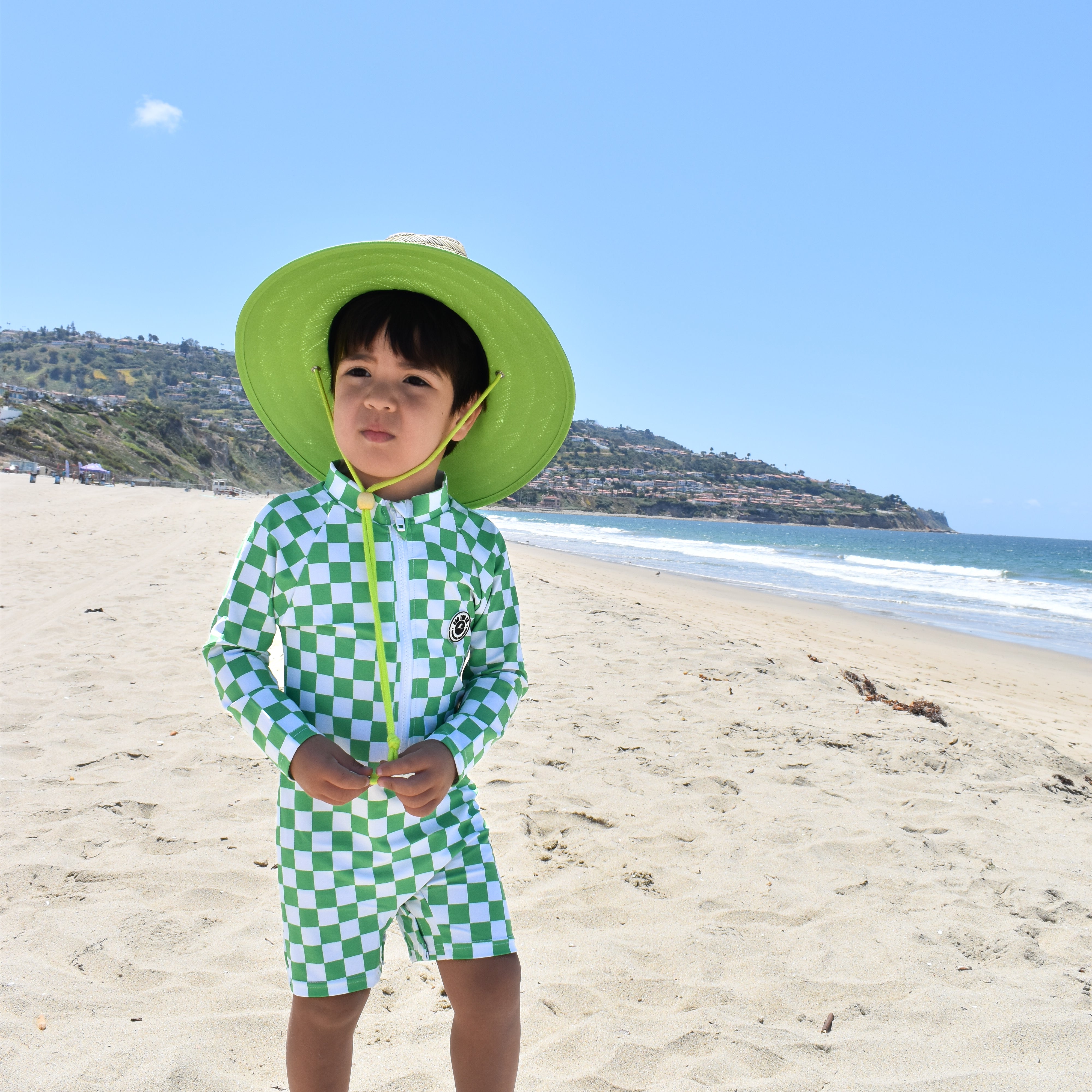 Child in green checkered swimsuit and hat standing on a sandy beach with ocean and clear sky in the background.