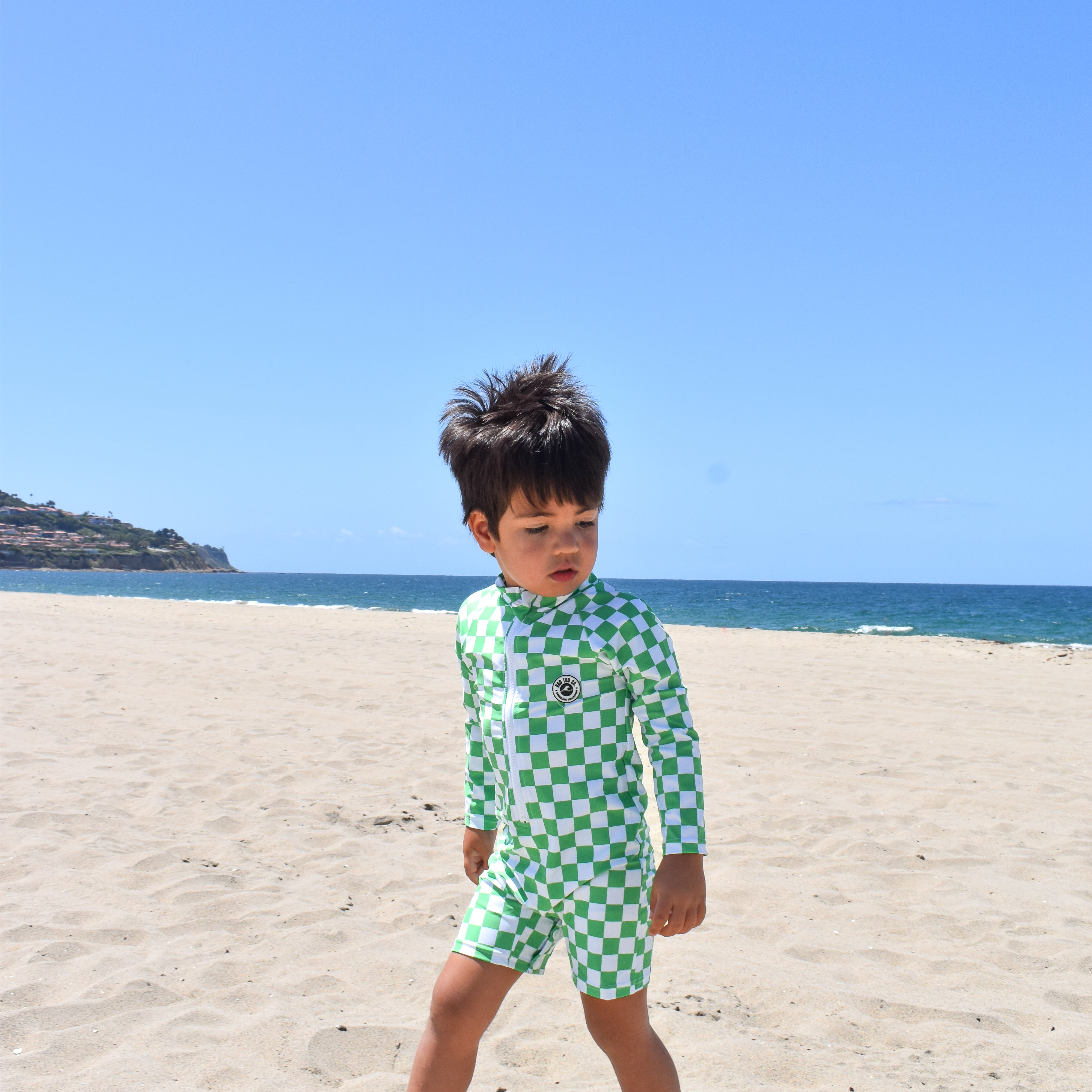 Child in a green checkered swimsuit standing on a sandy beach with clear blue sky.