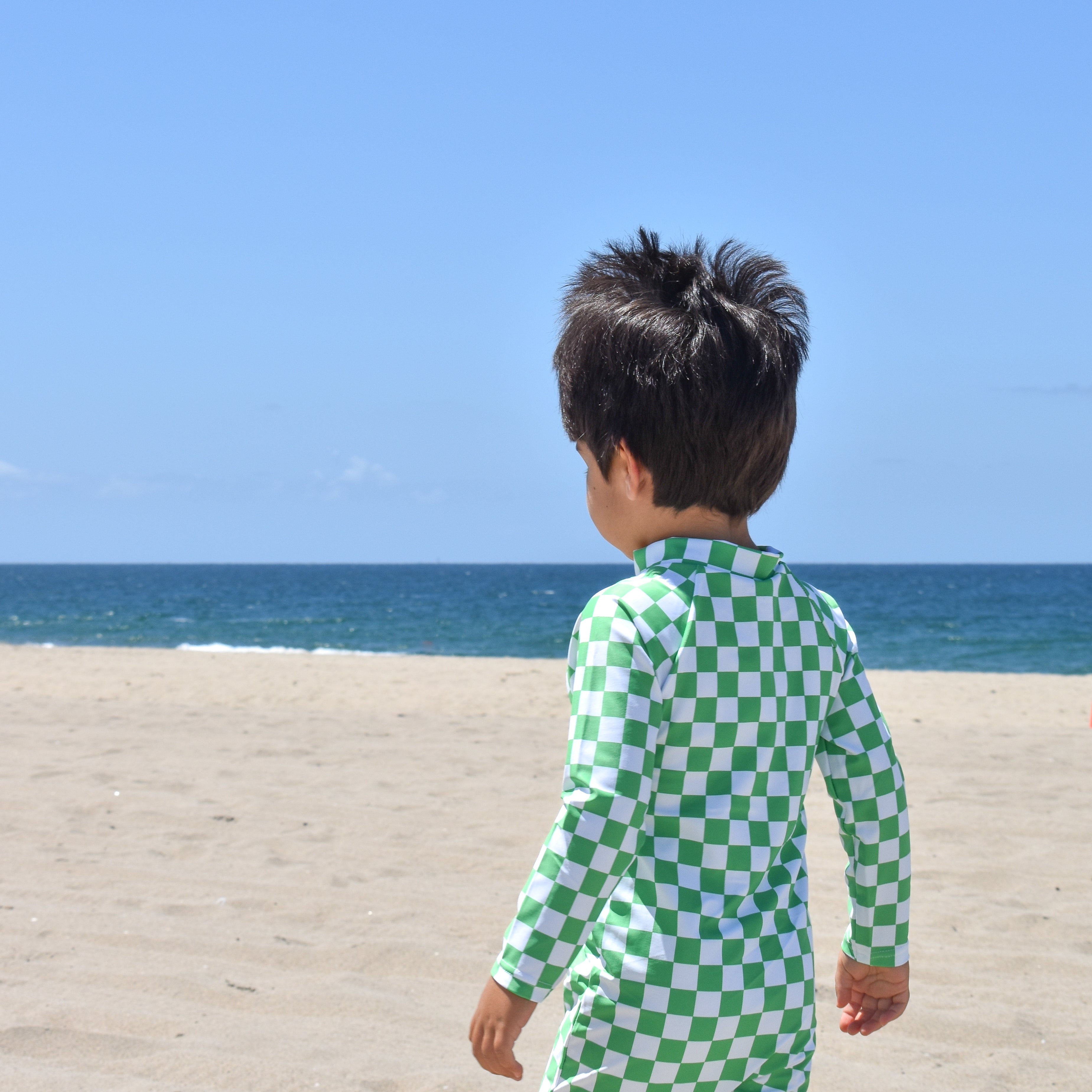 Child wearing a green checkered swimsuit standing on a sandy beach with ocean in the background