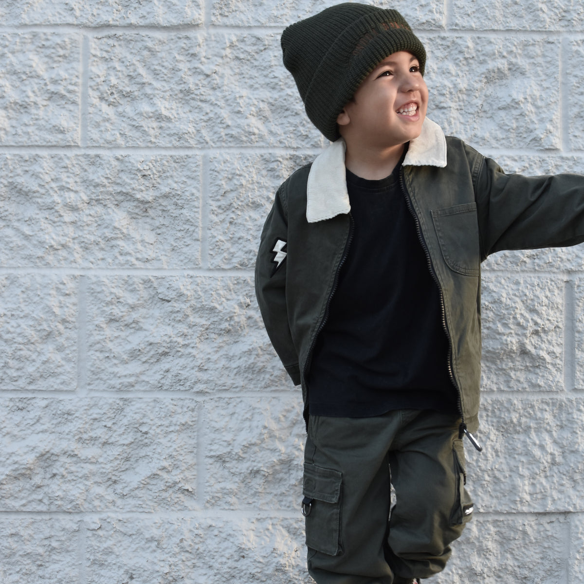 A young boy wearing a dark olive green jacket with a white shearling collar, matching cargo pants, a black t-shirt, and a dark knit beanie, posing against a light gray textured brick wall.