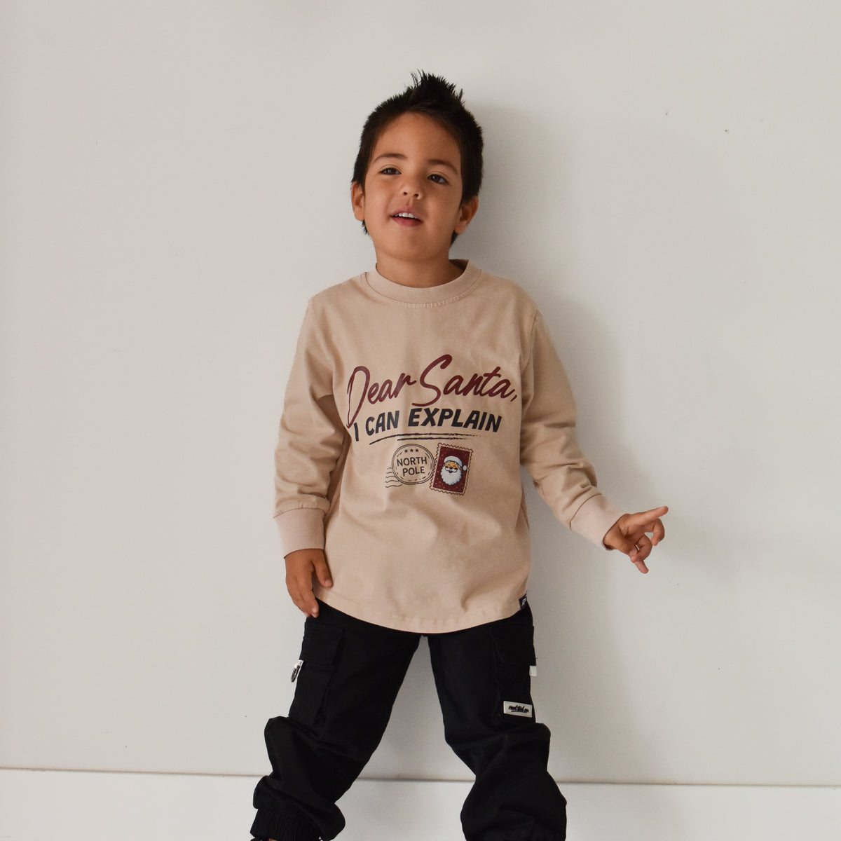 A young boy wearing a beige long-sleeve graphic sweatshirt and black cargo pants, standing against a plain white wall.