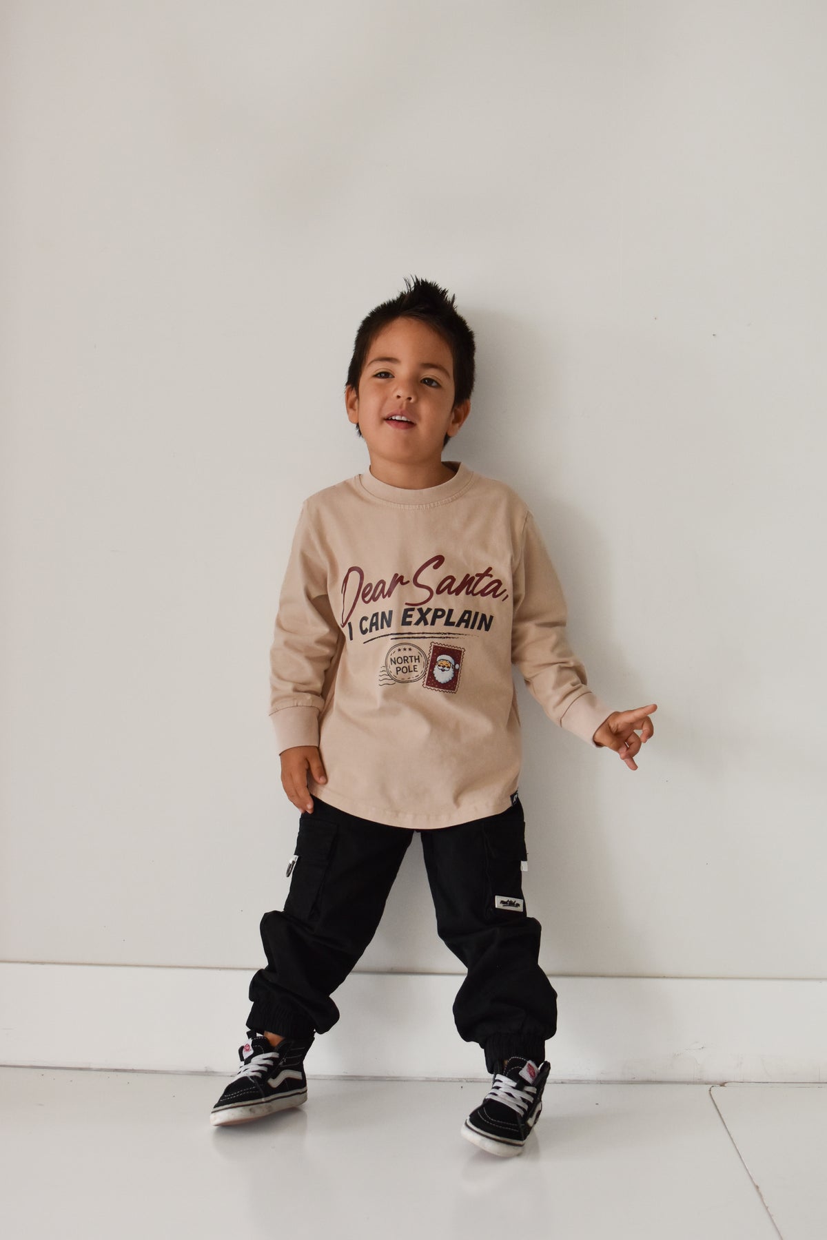 A young boy wearing a beige long-sleeve graphic sweatshirt and black cargo pants, standing against a plain white wall.