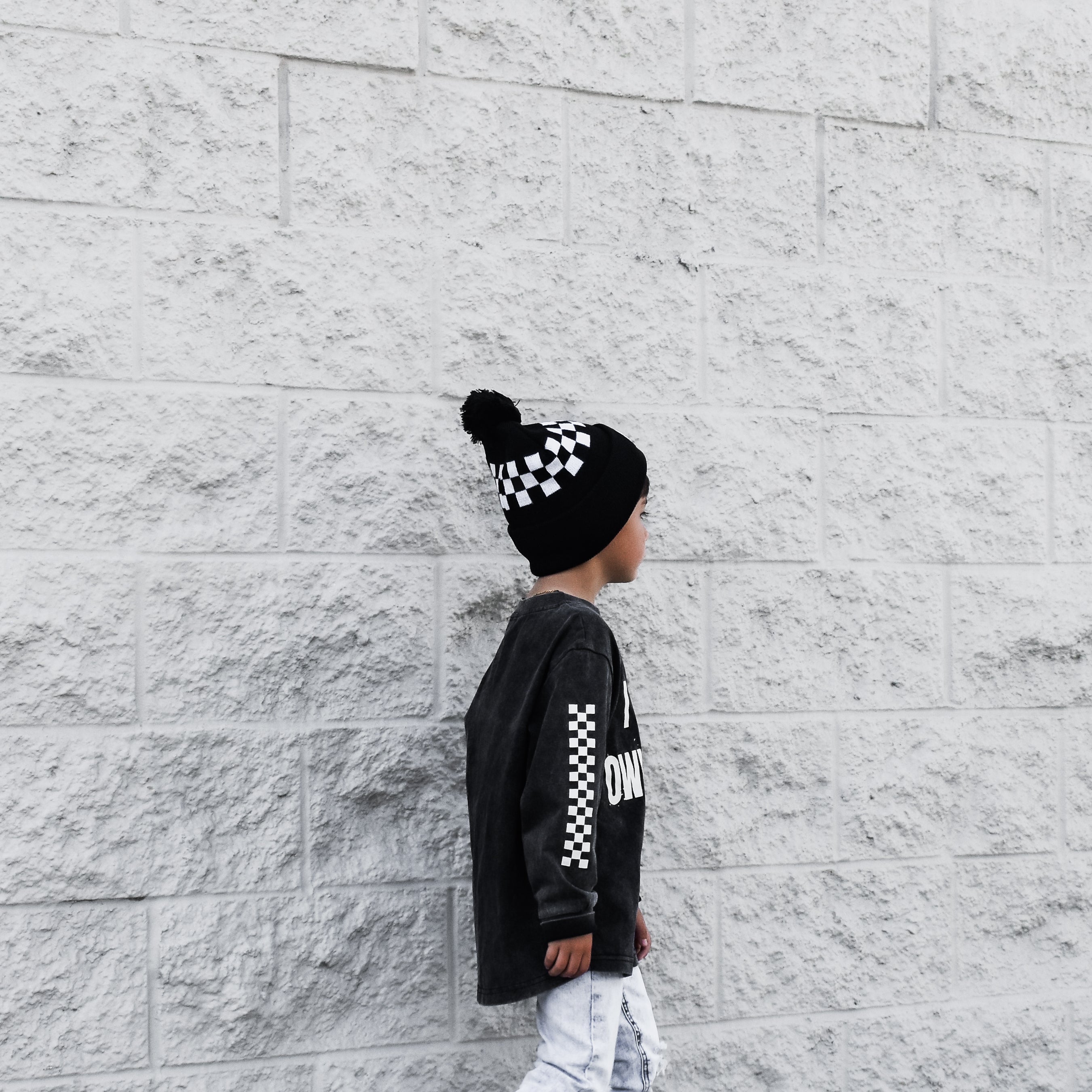 Child wearing a black long-sleeve shirt with white text, white pants, and a checkered beanie against a light gray brick wall.