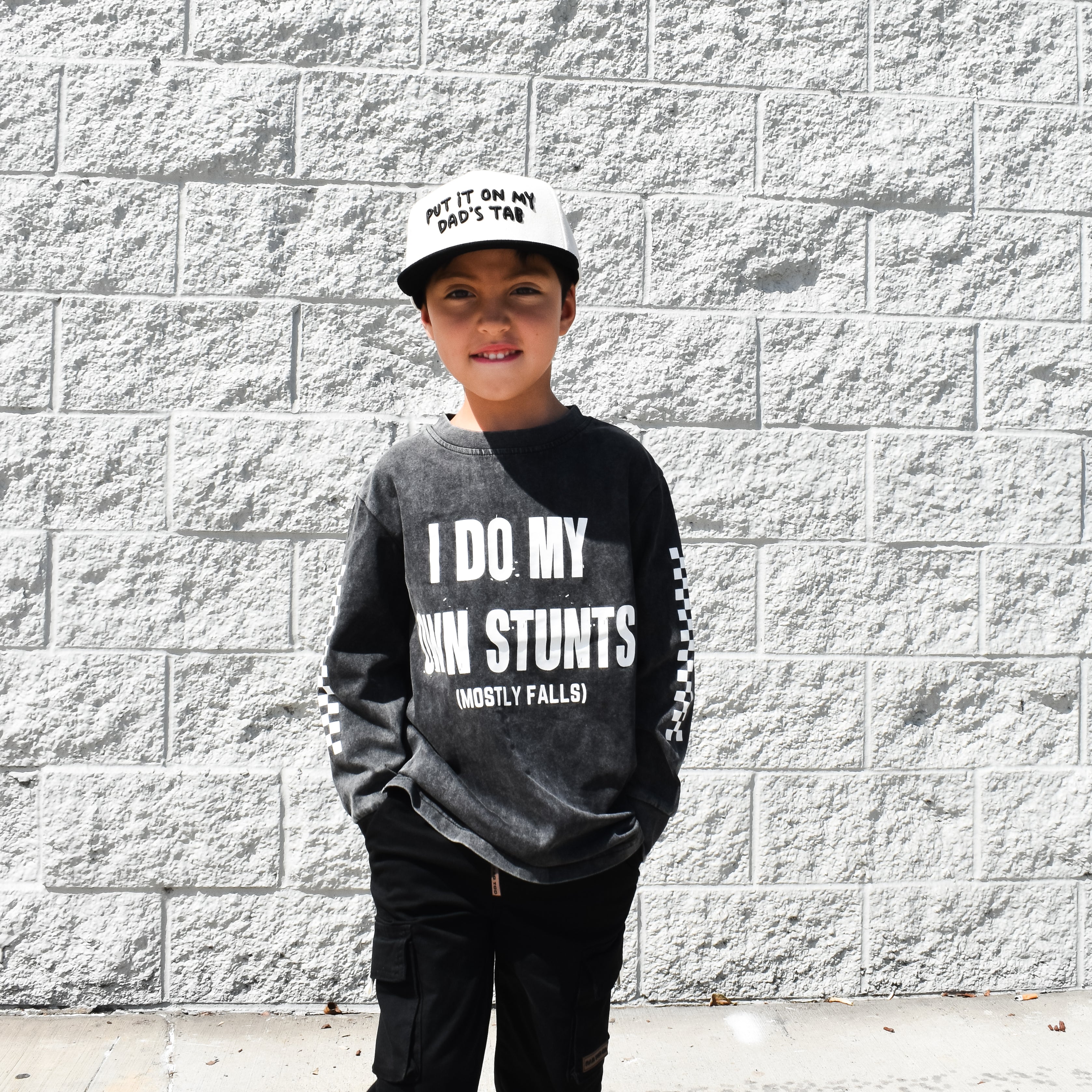Child wearing a black sweatshirt with text, standing against a gray brick wall.