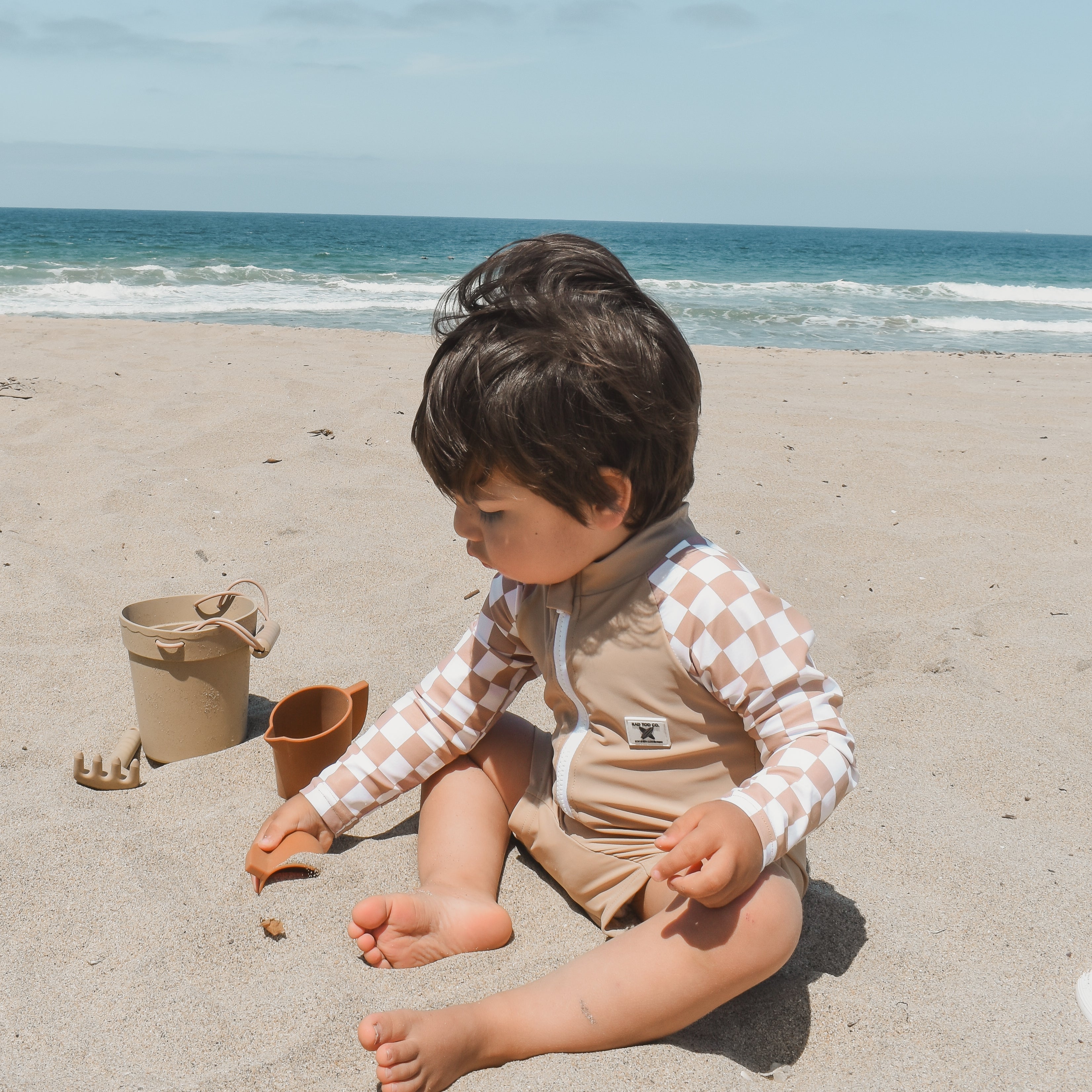 Toddler wearing the Dune Checkers one-piece rashguard with beige body and white checker sleeves, playing in the sand.