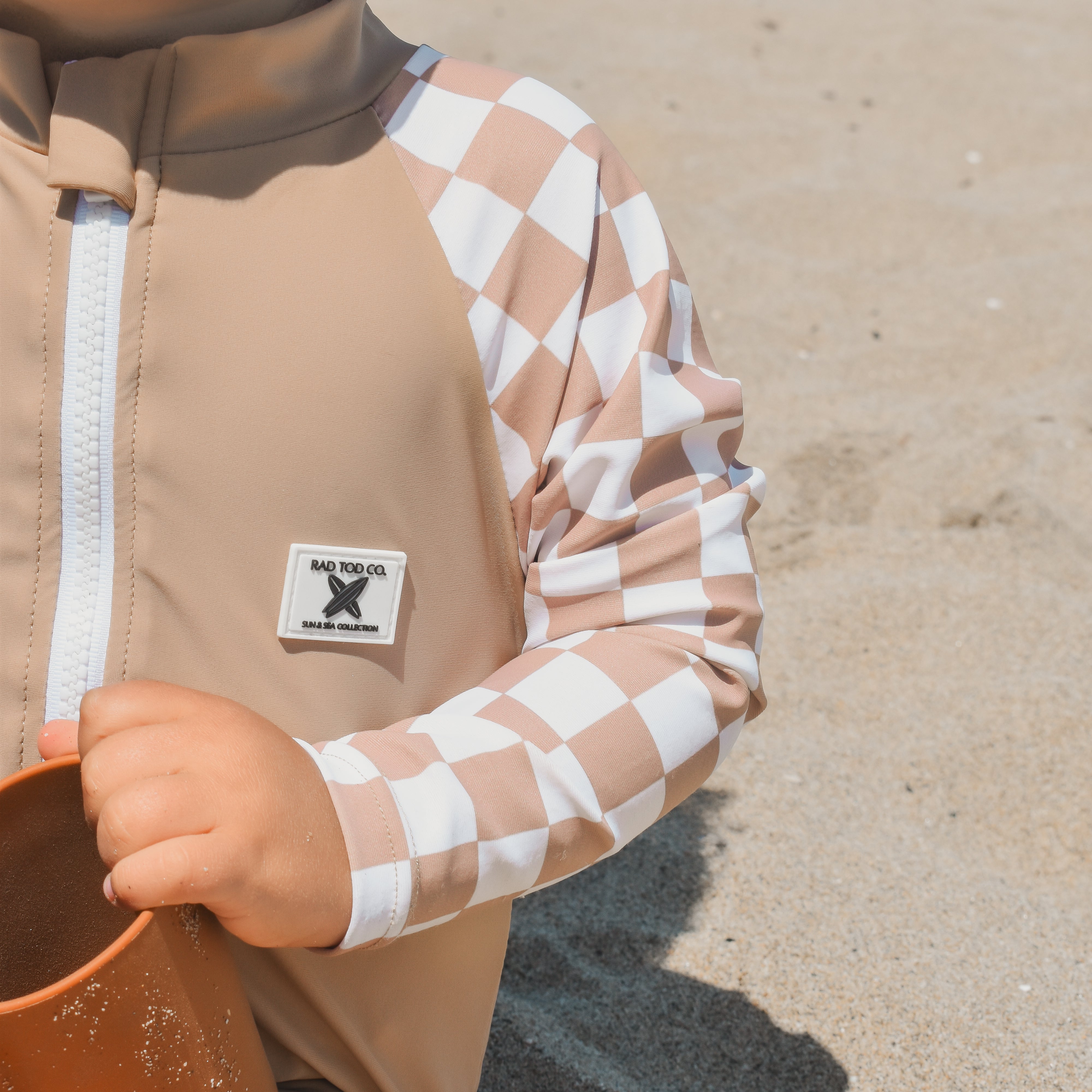 Toddler wearing the Dune Checkers one-piece rashguard with beige body and white checker sleeves, sitting on the beach.