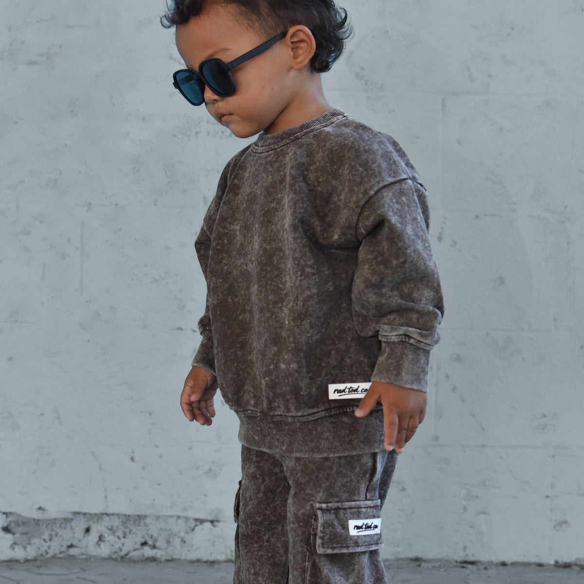 A toddler wearing a matching washed dark brown sweatshirt and cargo pants set, styled with black sunglasses and white sneakers, standing against a light gray concrete wall.