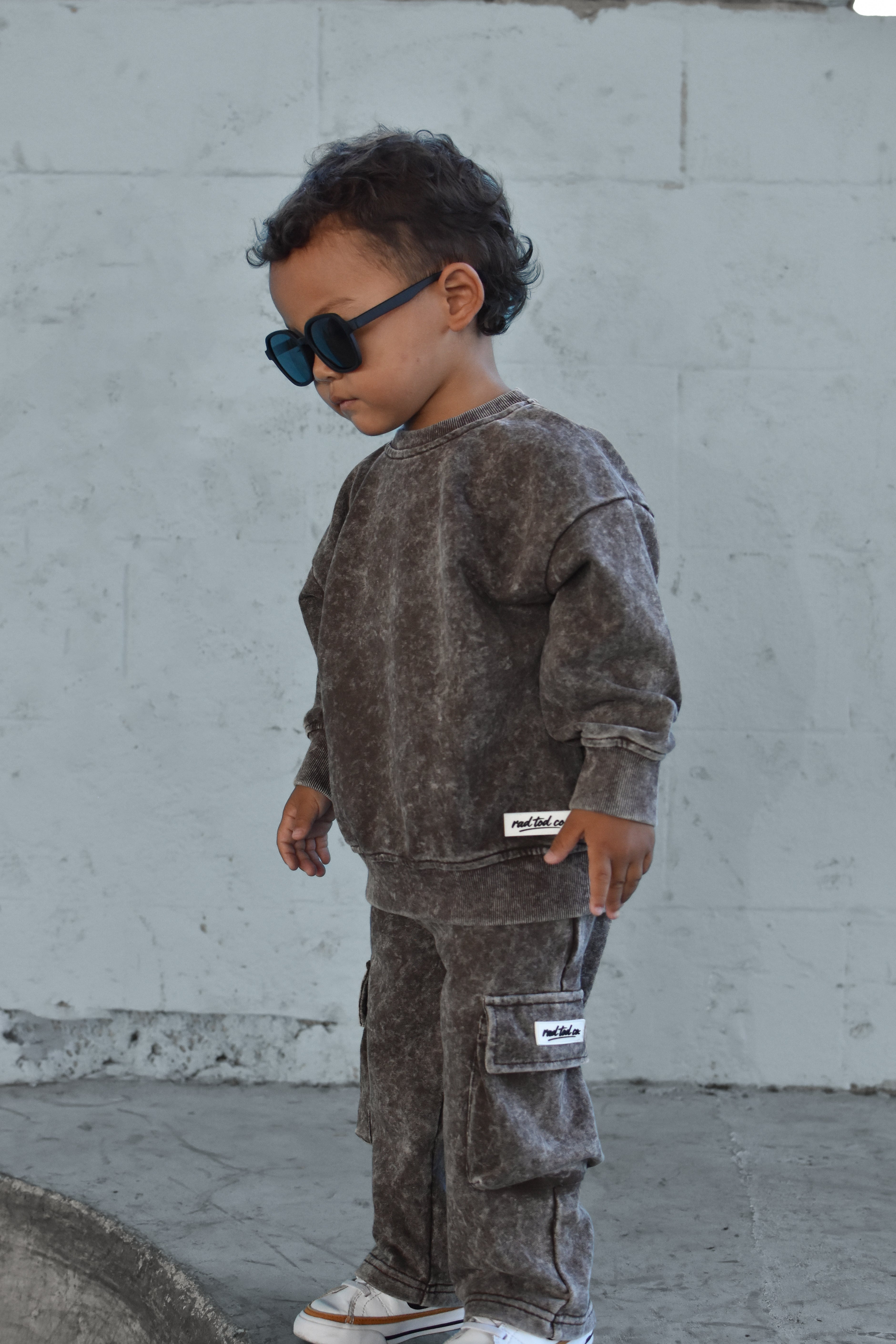 A toddler wearing a matching washed dark brown sweatshirt and cargo pants set, styled with black sunglasses and white sneakers, standing against a light gray concrete wall.