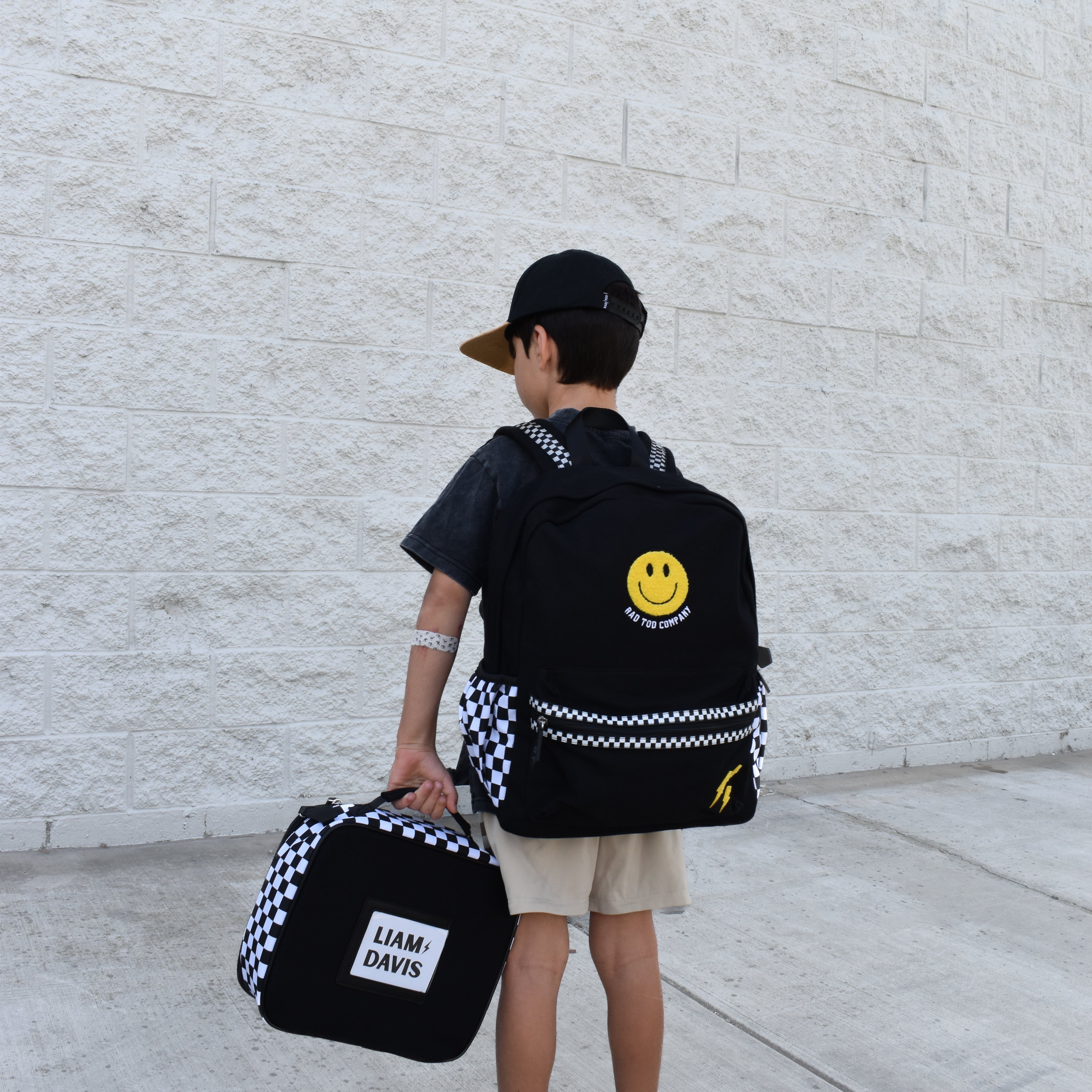 Child holding a black backpack with a smiley face design and a smaller bag, standing against a light gray wall.