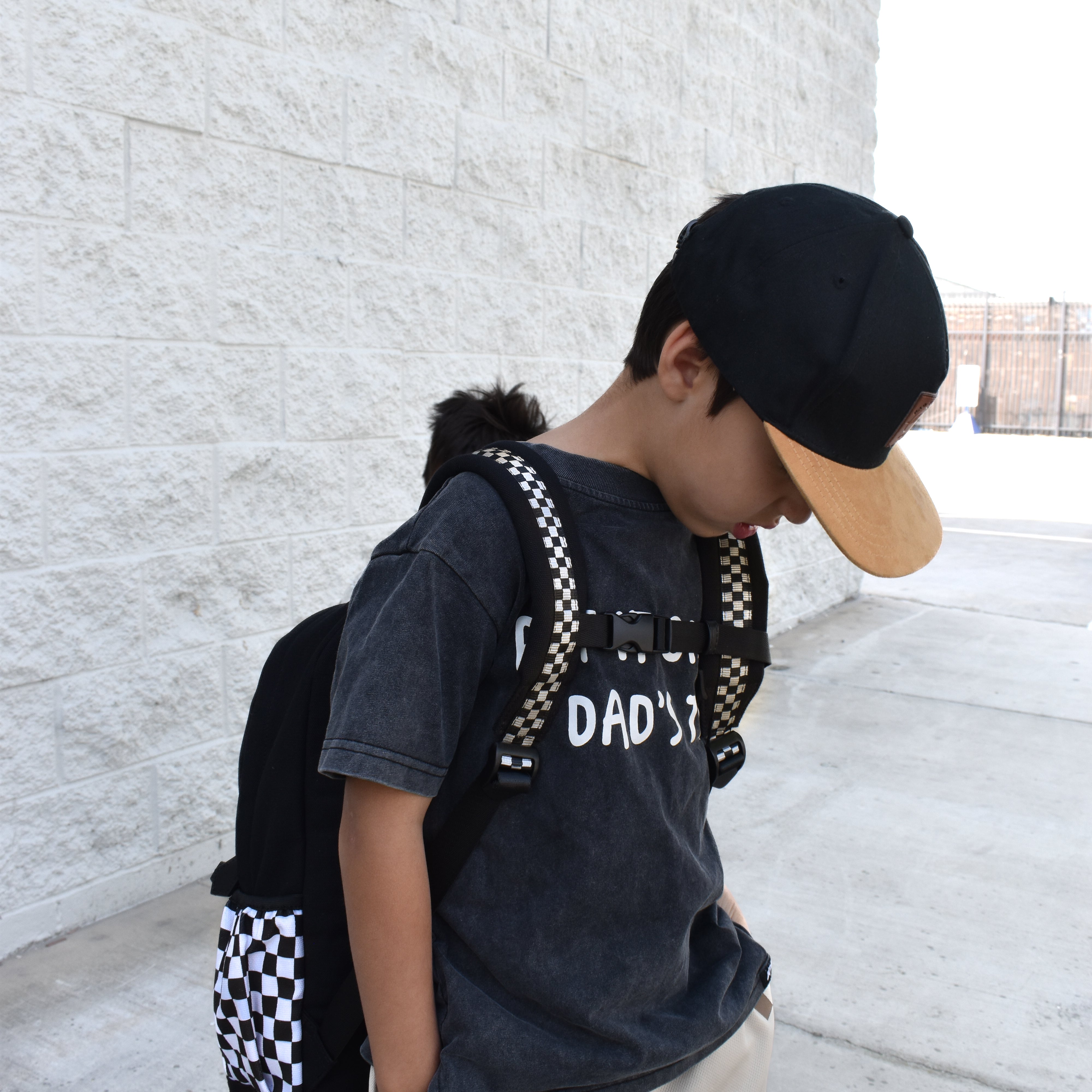 Child wearing a black t-shirt with text, beige shorts, and a checkered backpack against a light stone wall.