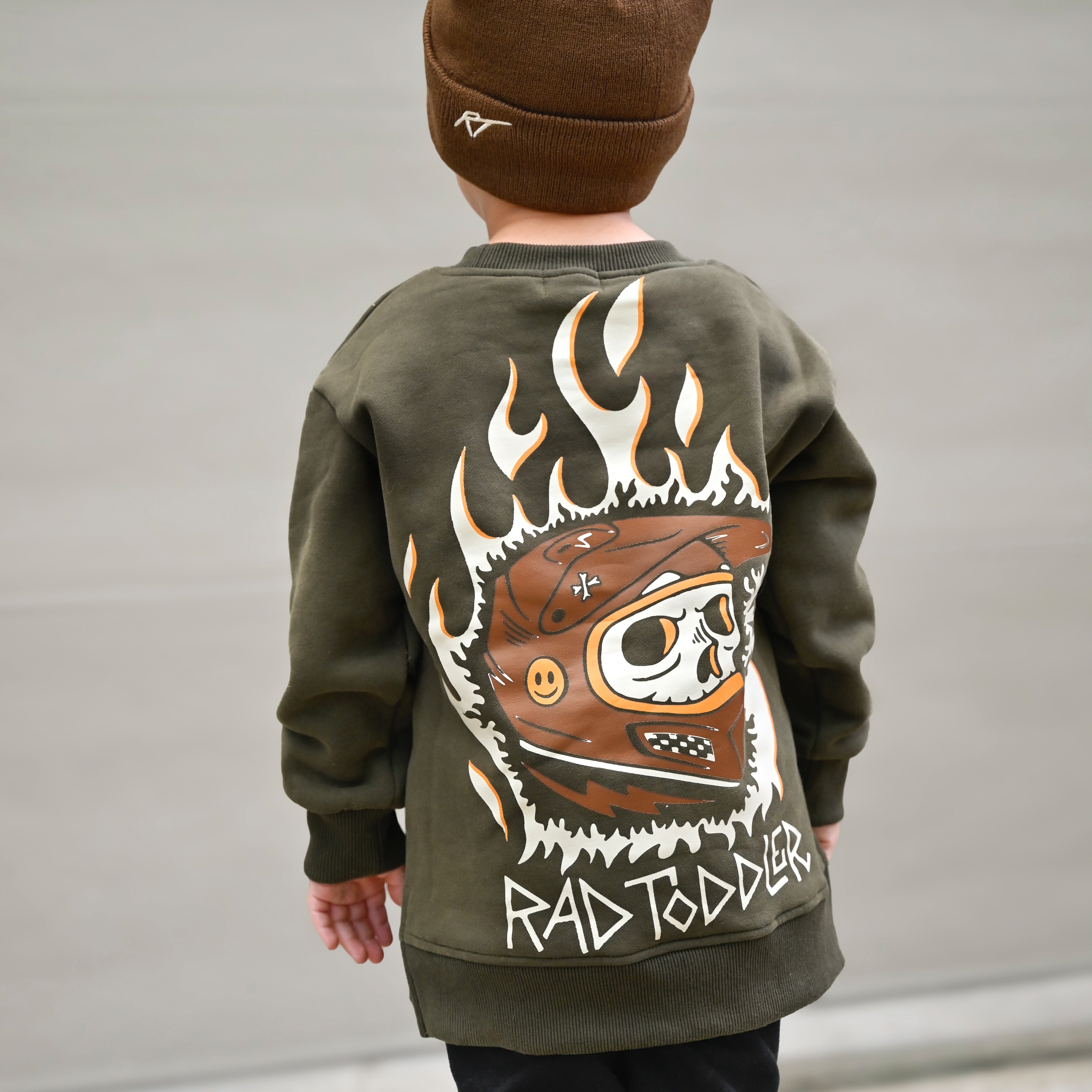Child wearing a green sweatshirt with a graphic design and brown beanie, standing against a plain background.