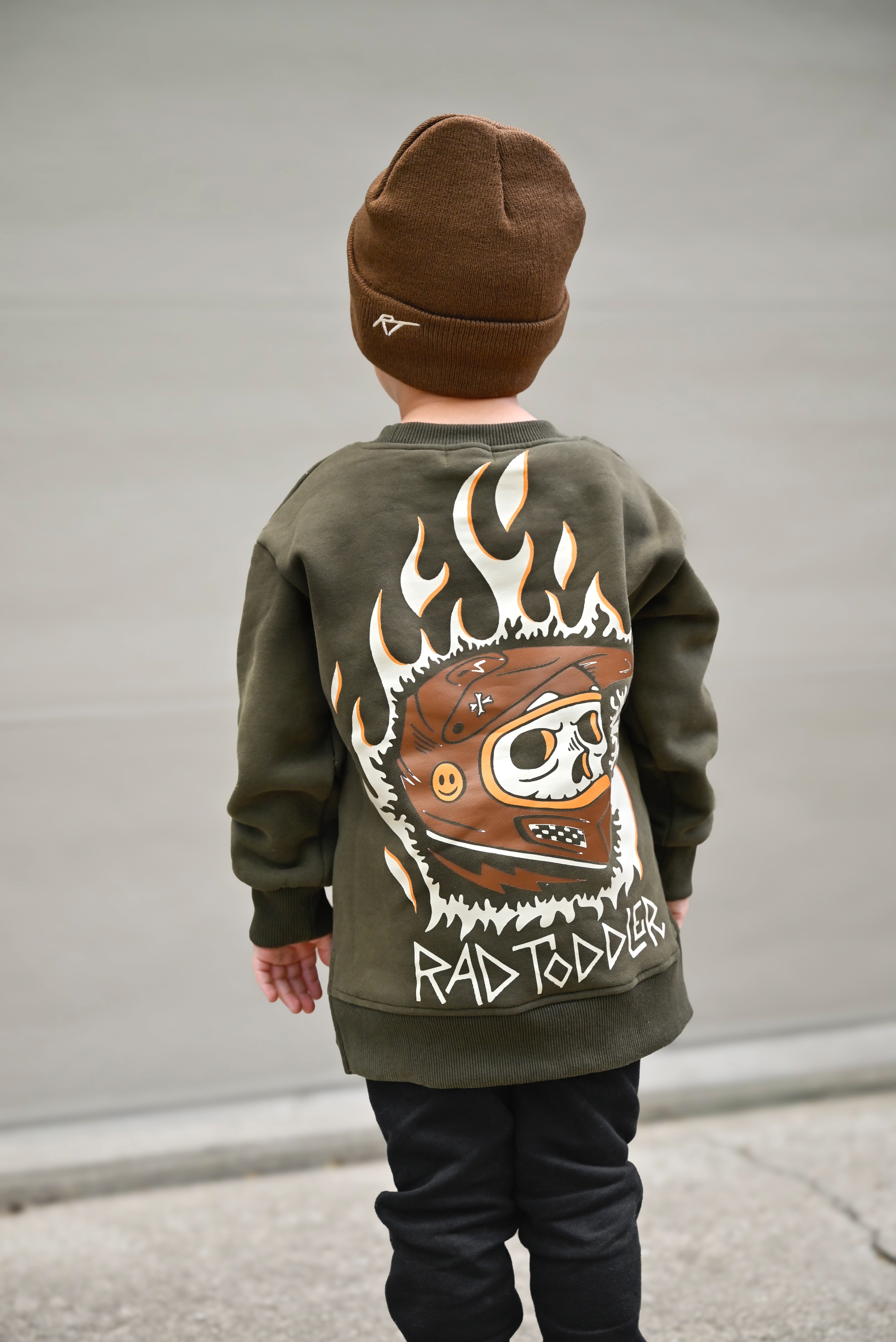 Child wearing a green sweatshirt with a graphic design and brown beanie, standing against a plain background.