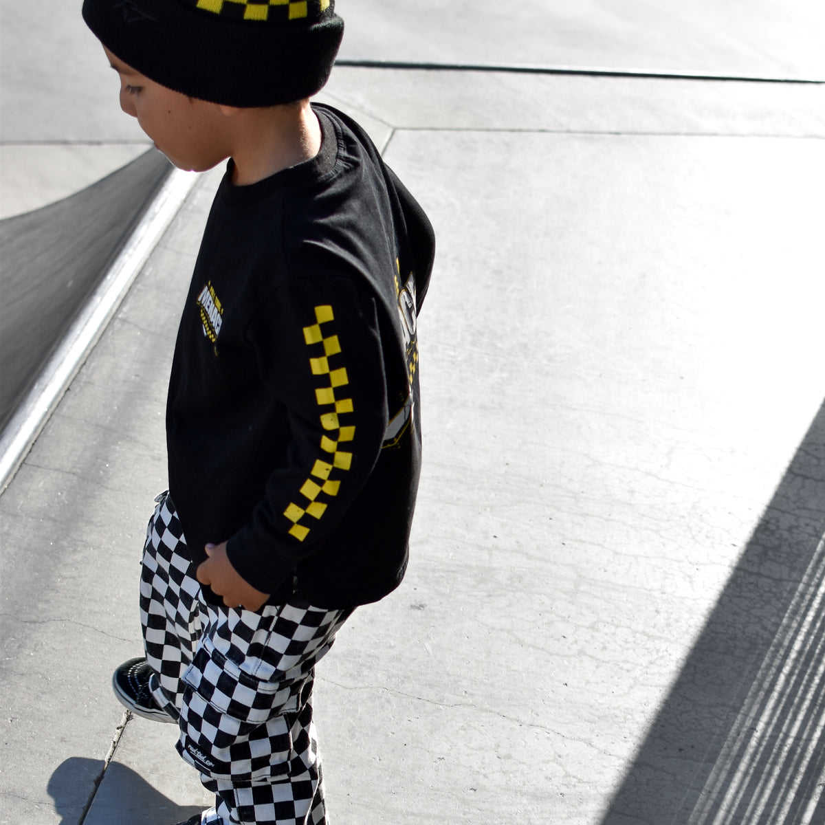 A toddler standing outdoors on a concrete surface, wearing a black beanie with yellow checkered accents, a black long-sleeve shirt with yellow checkered sleeve details, and black-and-white checkered pants.
