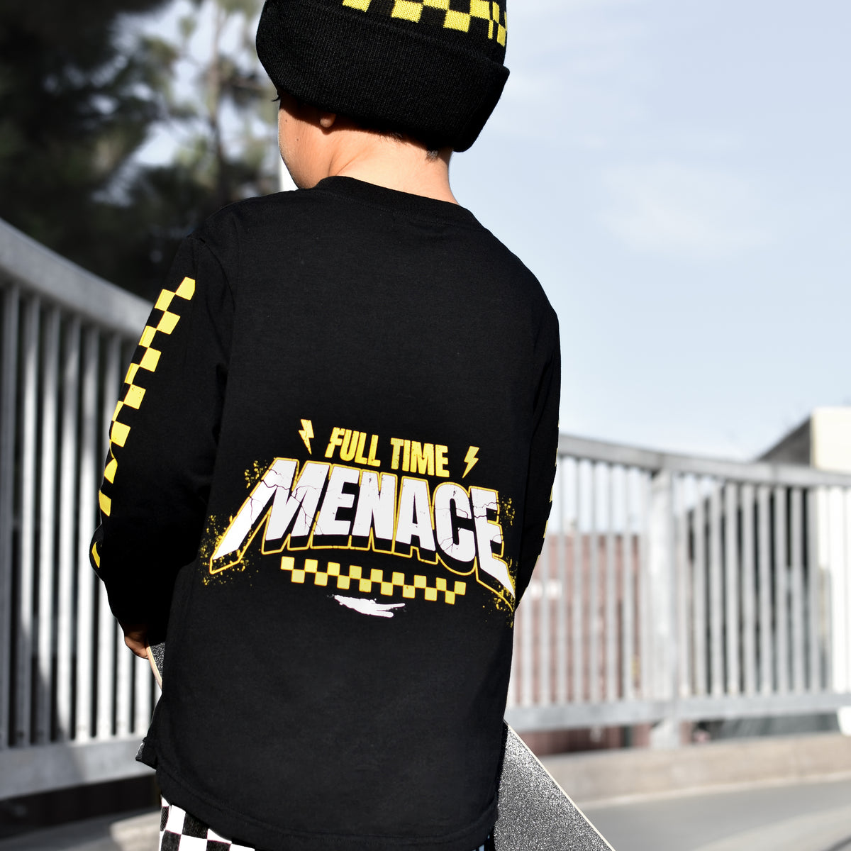 A child viewed from behind wearing a black long-sleeve t-shirt with a yellow and white 'Full Time Menace' graphic on the back, a matching black and yellow beanie, and black-and-white checkered pants, standing outdoors in a skatepark.