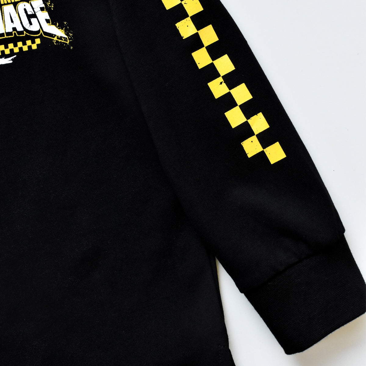 Close-up of a black long-sleeve shirt with a yellow and black checkered pattern down one sleeve and a partial graphic on the chest, shown on a light gray background.