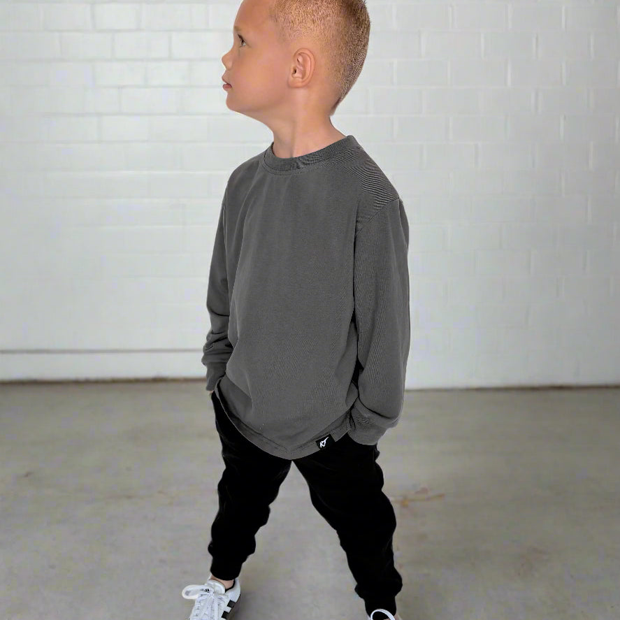 Child wearing a gray sweatshirt and black pants standing against a white brick wall.