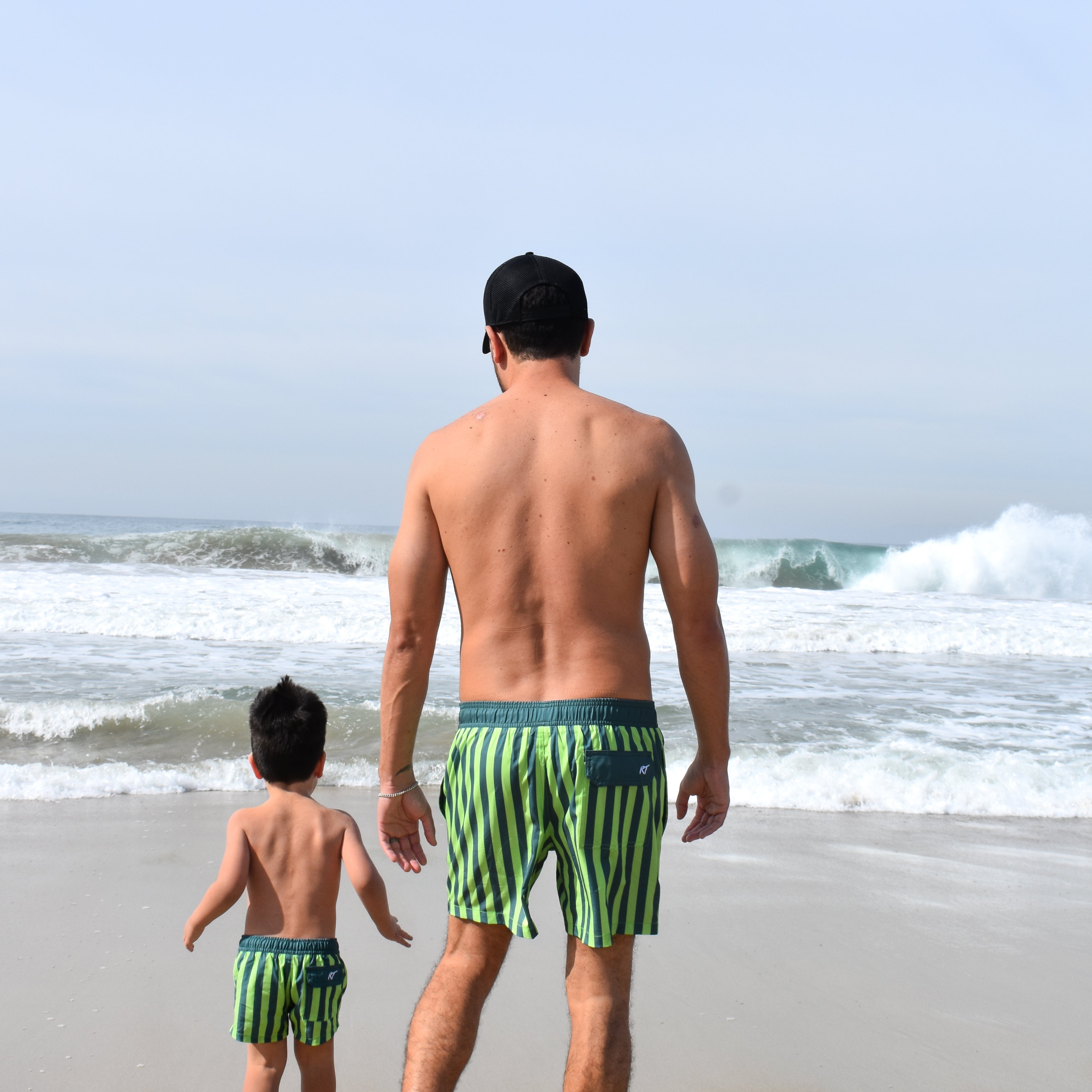 Man and child walking on a beach wearing matching lime green and dark green striped swim trunks by rad toddler