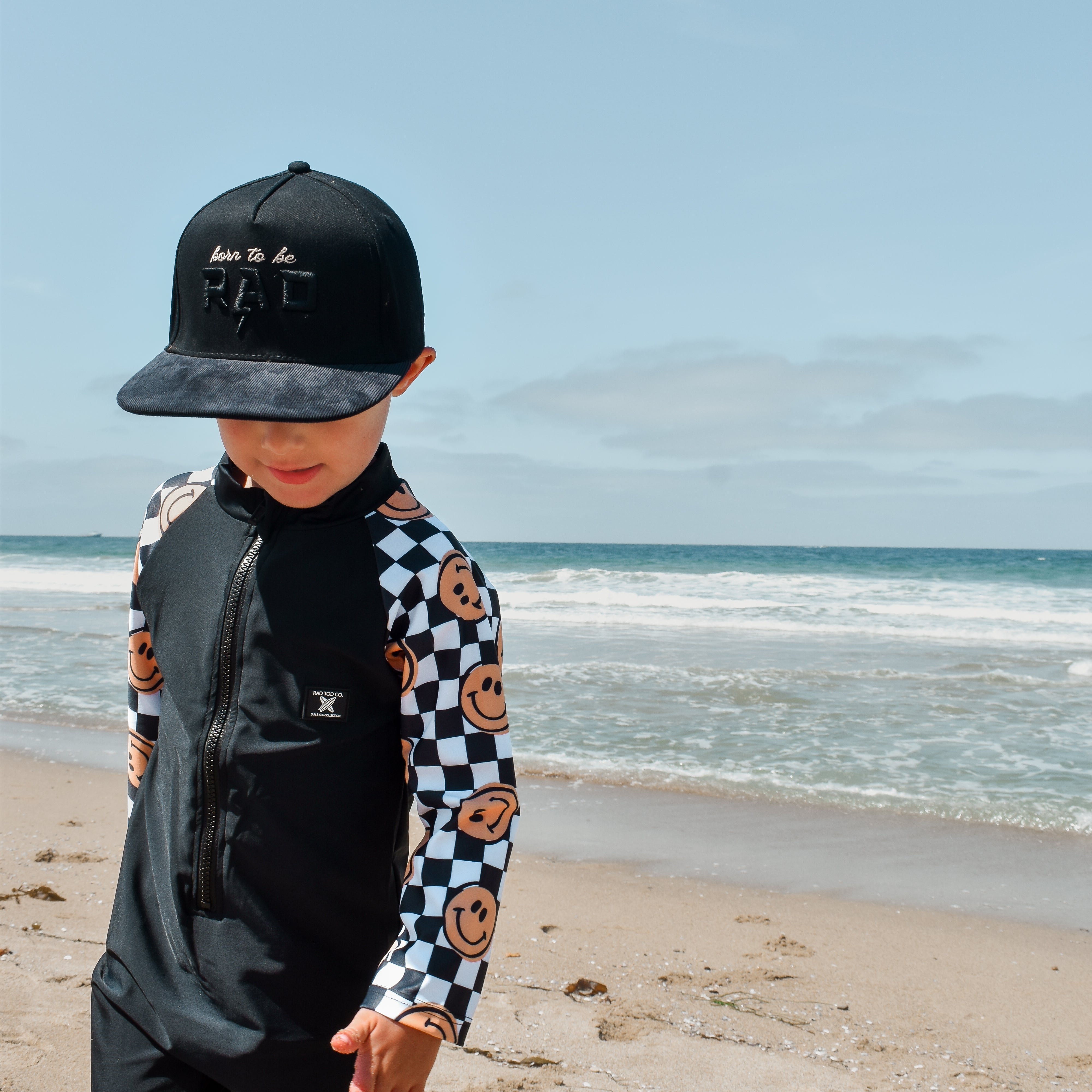 rad toddler boy wearing a checker-patterned one-piece rashguard with smiley faces on the sleeves, standing on the beach near the shoreline