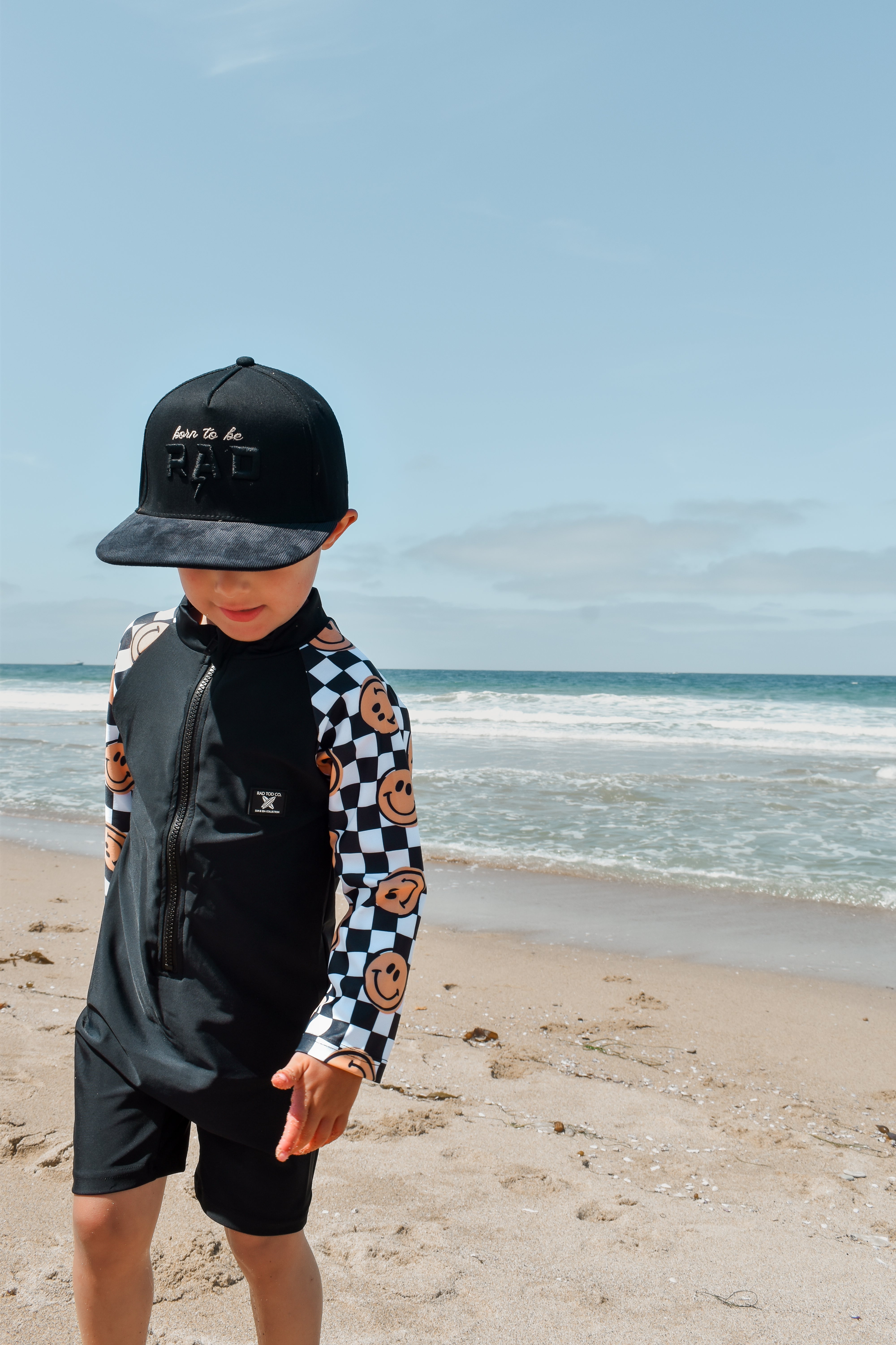 rad toddler boy wearing a checker-patterned one-piece rashguard with smiley faces on the sleeves, standing on the beach near the shoreline
