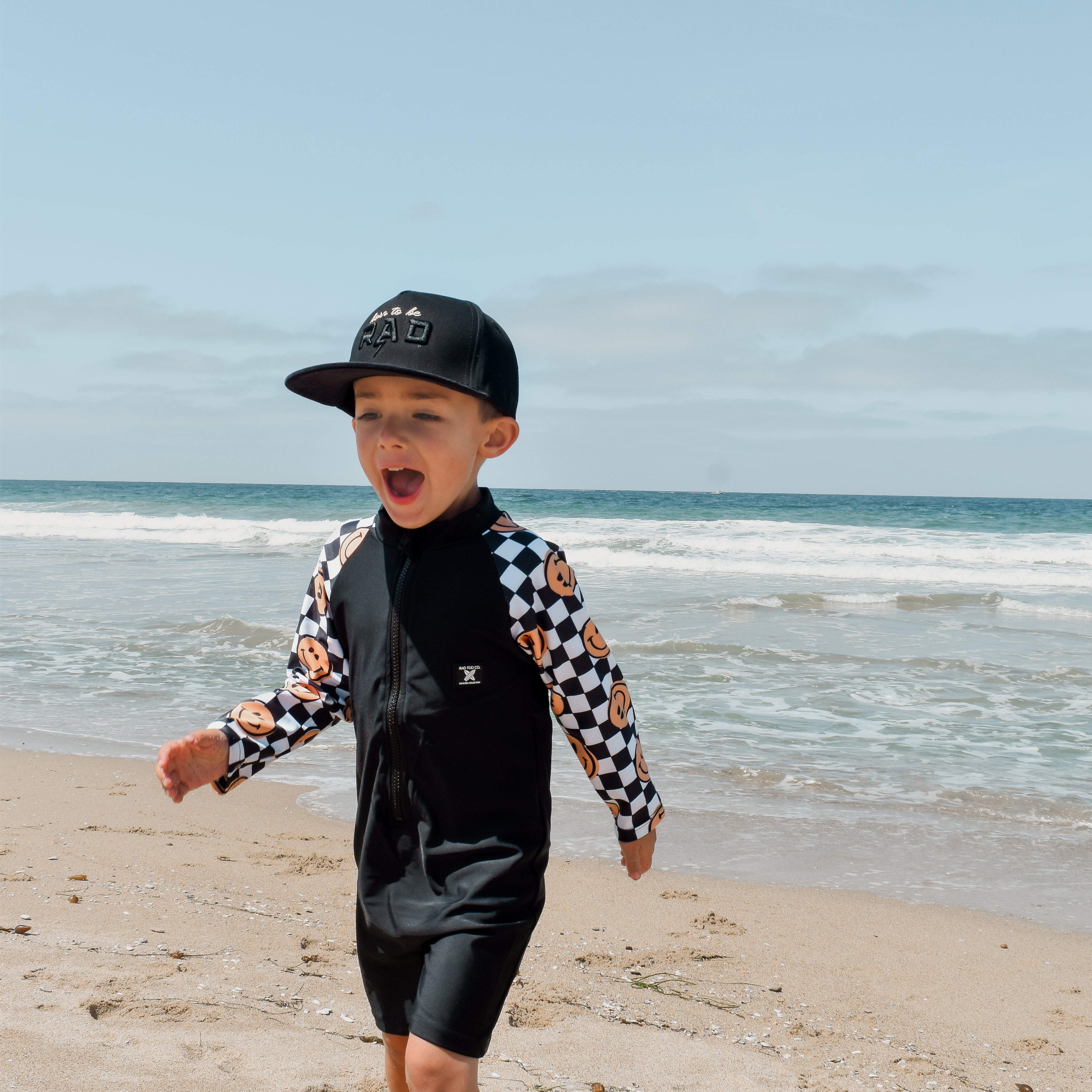 Young boy wearing a checker-patterned one-piece rashguard with smiley faces on the sleeves, standing on the beach near the shoreline