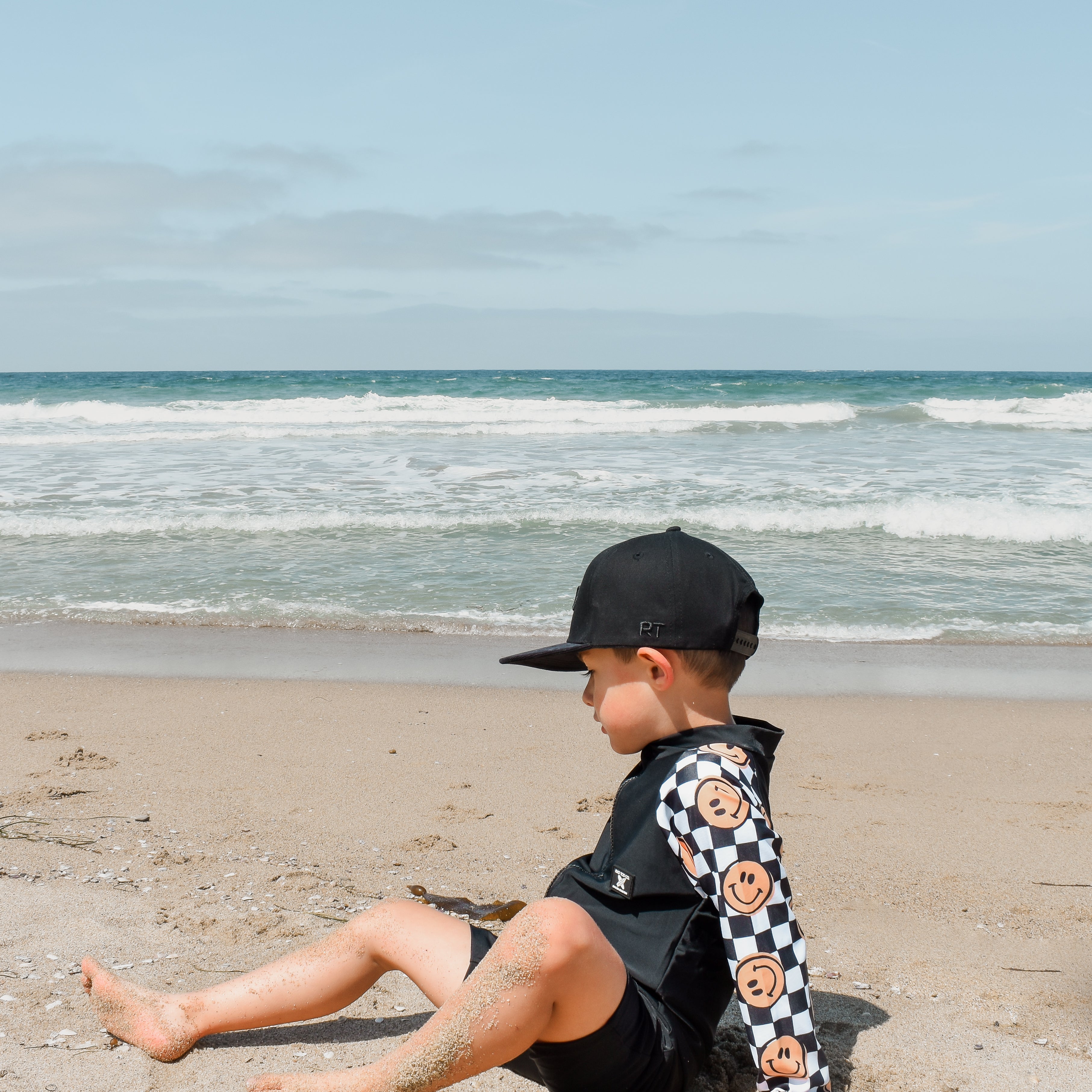 Young boy wearing a checker-patterned one-piece rashguard with smiley faces on the sleeves, relaxing on the beach near the shoreline