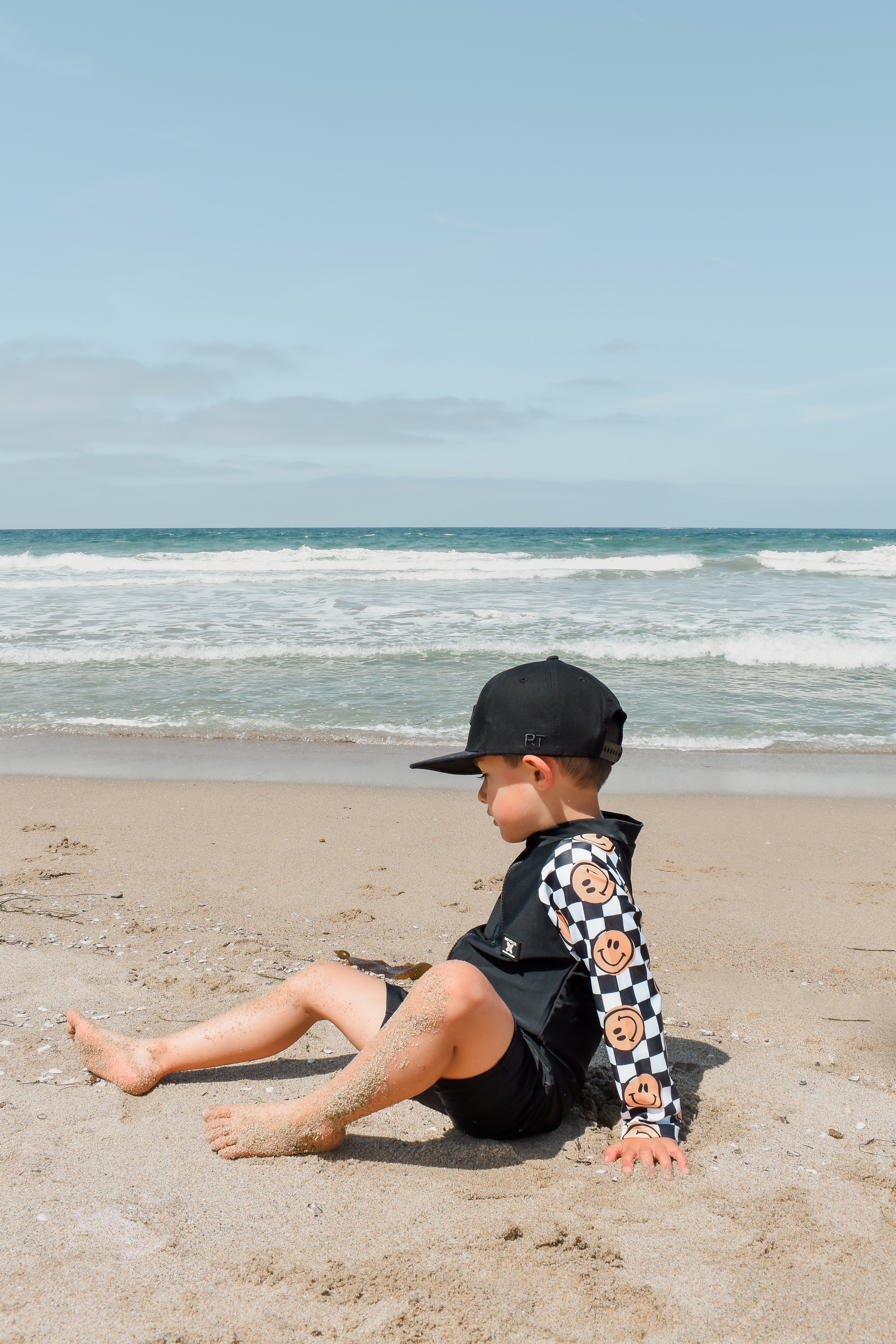 Young boy wearing a checker-patterned one-piece rashguard with smiley faces on the sleeves, relaxing on the beach near the shoreline