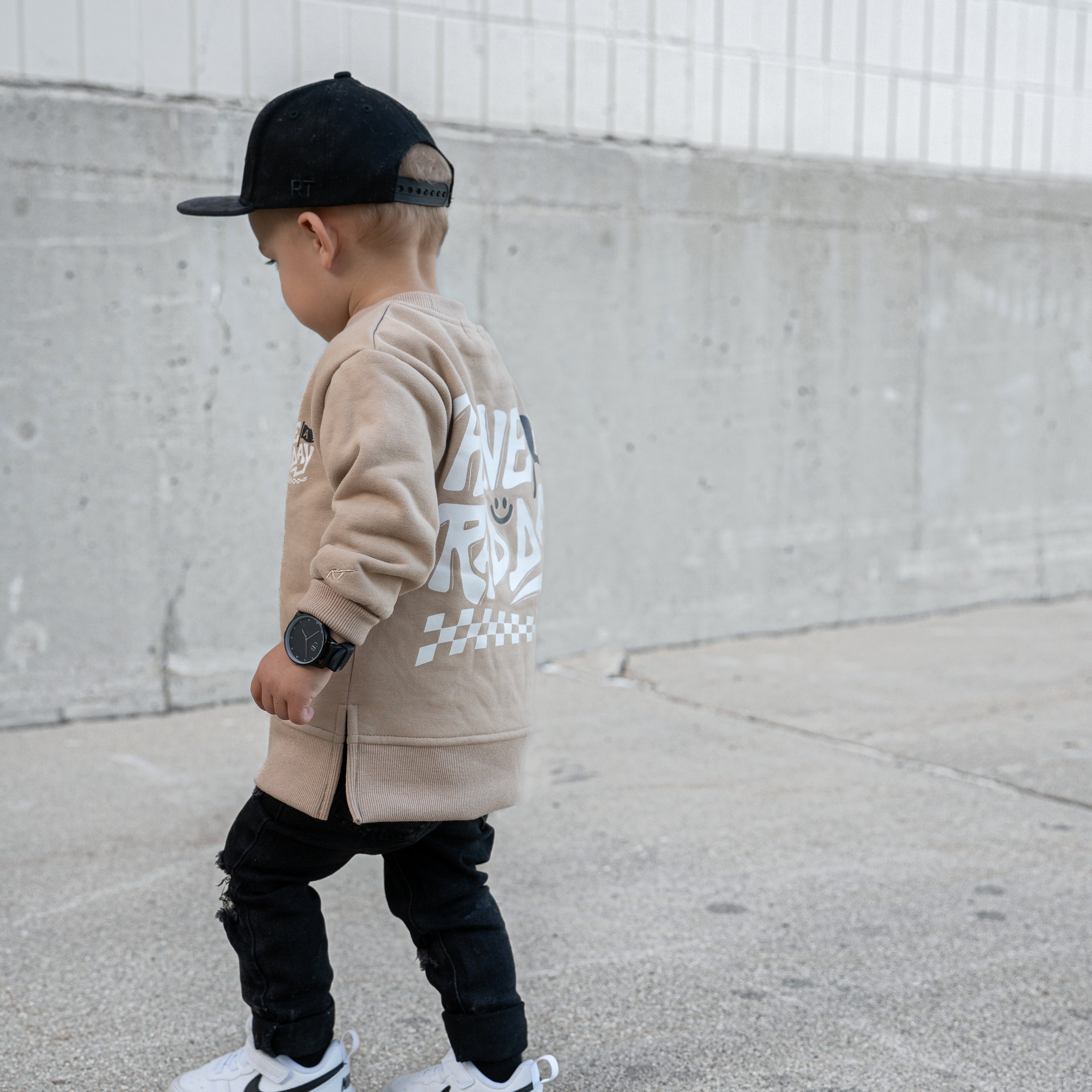 Child wearing a beige hoodie with text, black pants, and white sneakers against a concrete wall.