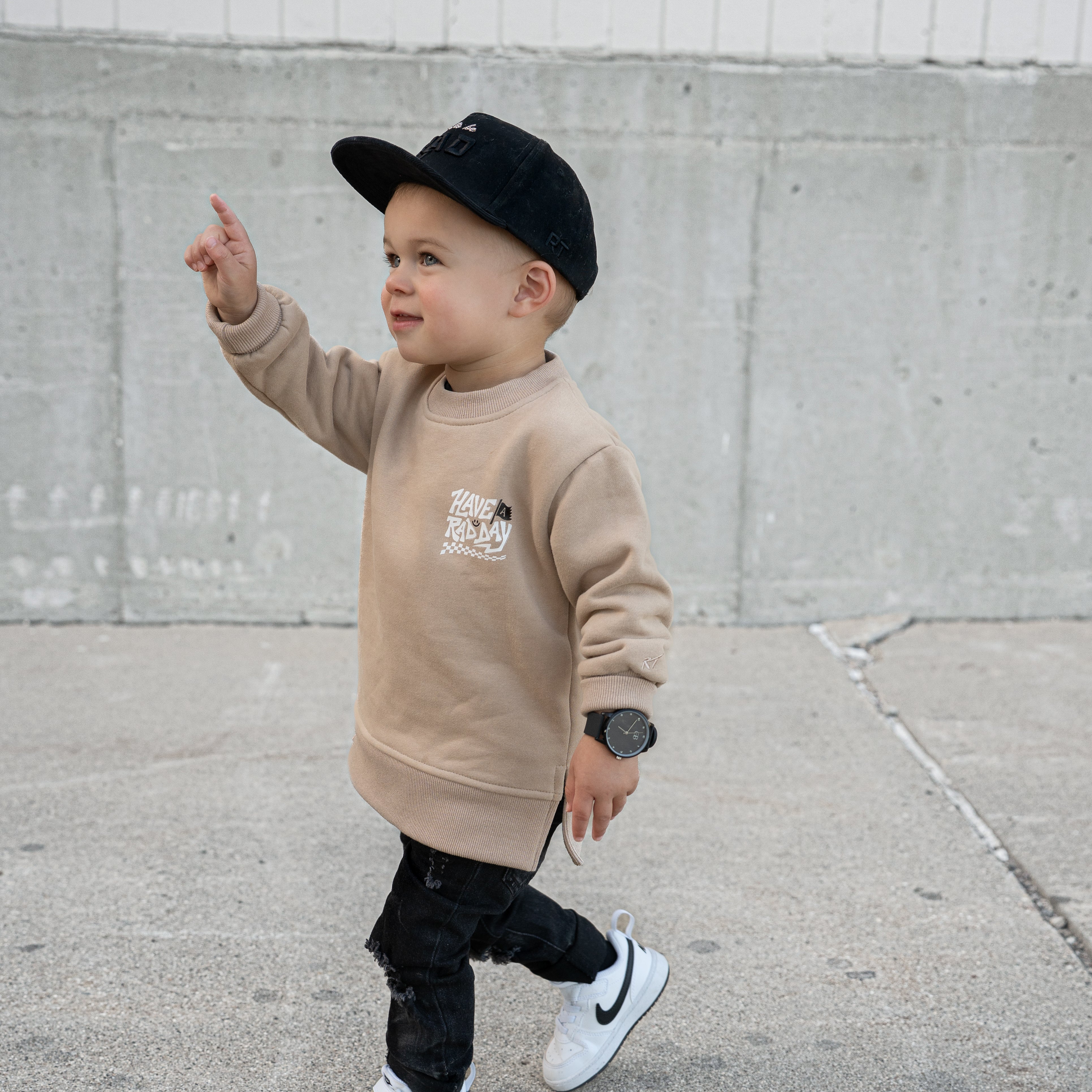 Child wearing a beige sweatshirt, black pants, and a black cap, standing on a sidewalk.