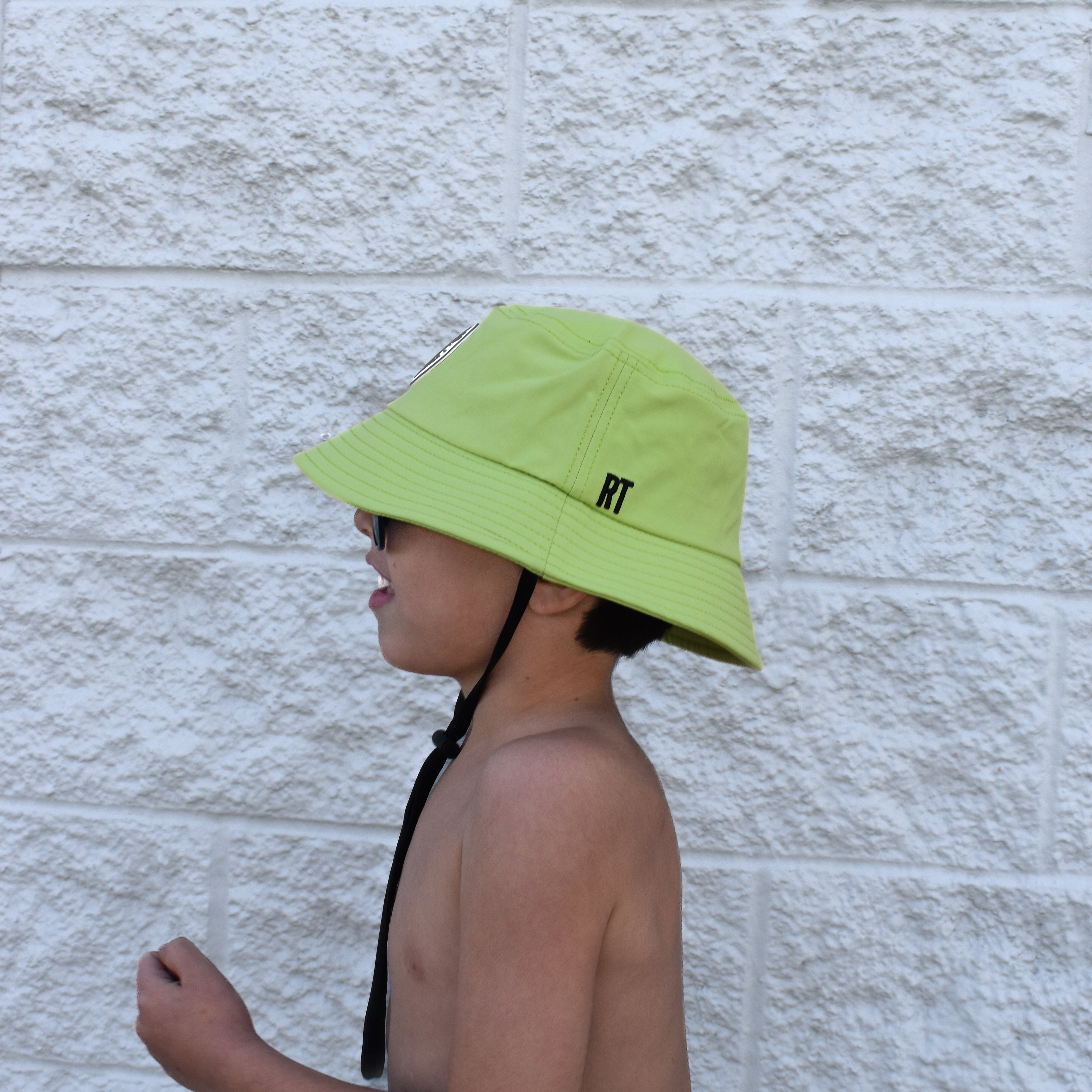 Child wearing a bright green bucket hat by Rad Toddler with a brand logo against a light stone wall.