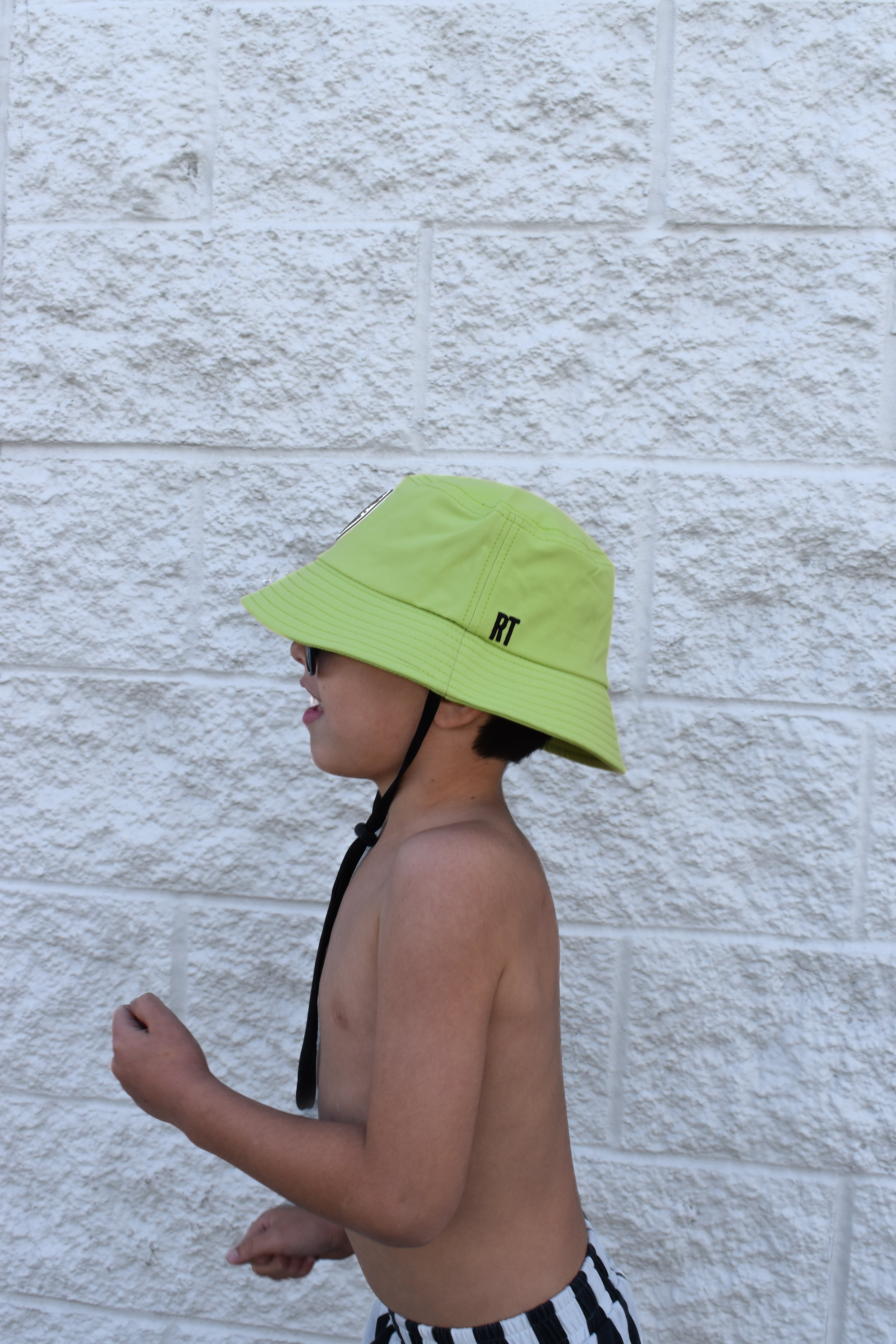 Child wearing a bright green bucket hat by Rad Toddler with a brand logo against a light stone wall.