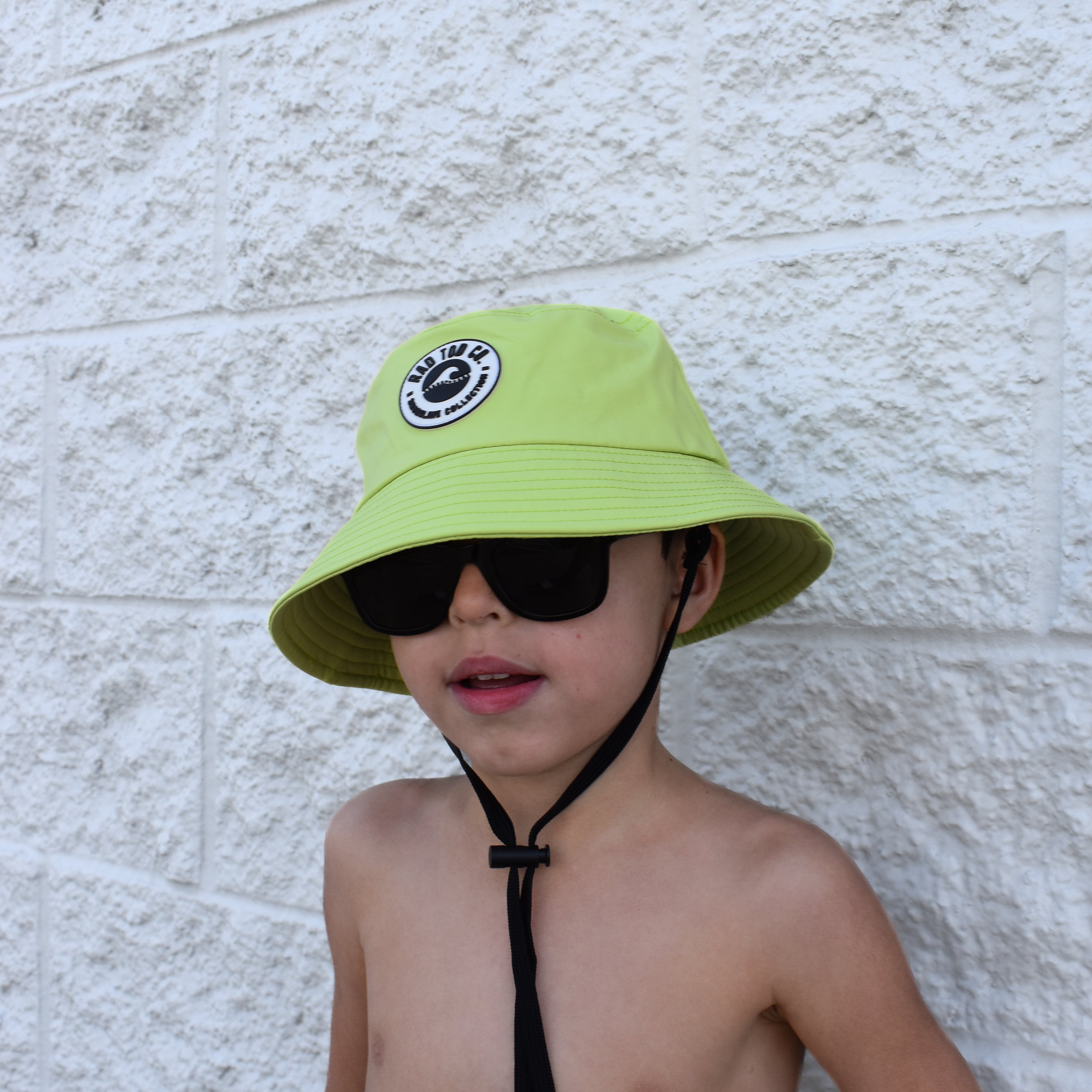 Child wearing a bright green bucket hat and sunglasses against a light stone wall.