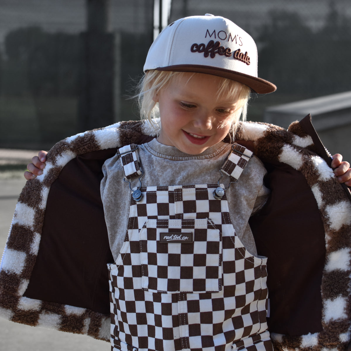 A young child wearing brown and white checkered overalls with a matching fuzzy checkered jacket draped open behind them, a gray shirt, and a white trucker hat that reads 'Moms coffee date'. The photo is taken outdoors on a paved surface with a fence and building in the background.