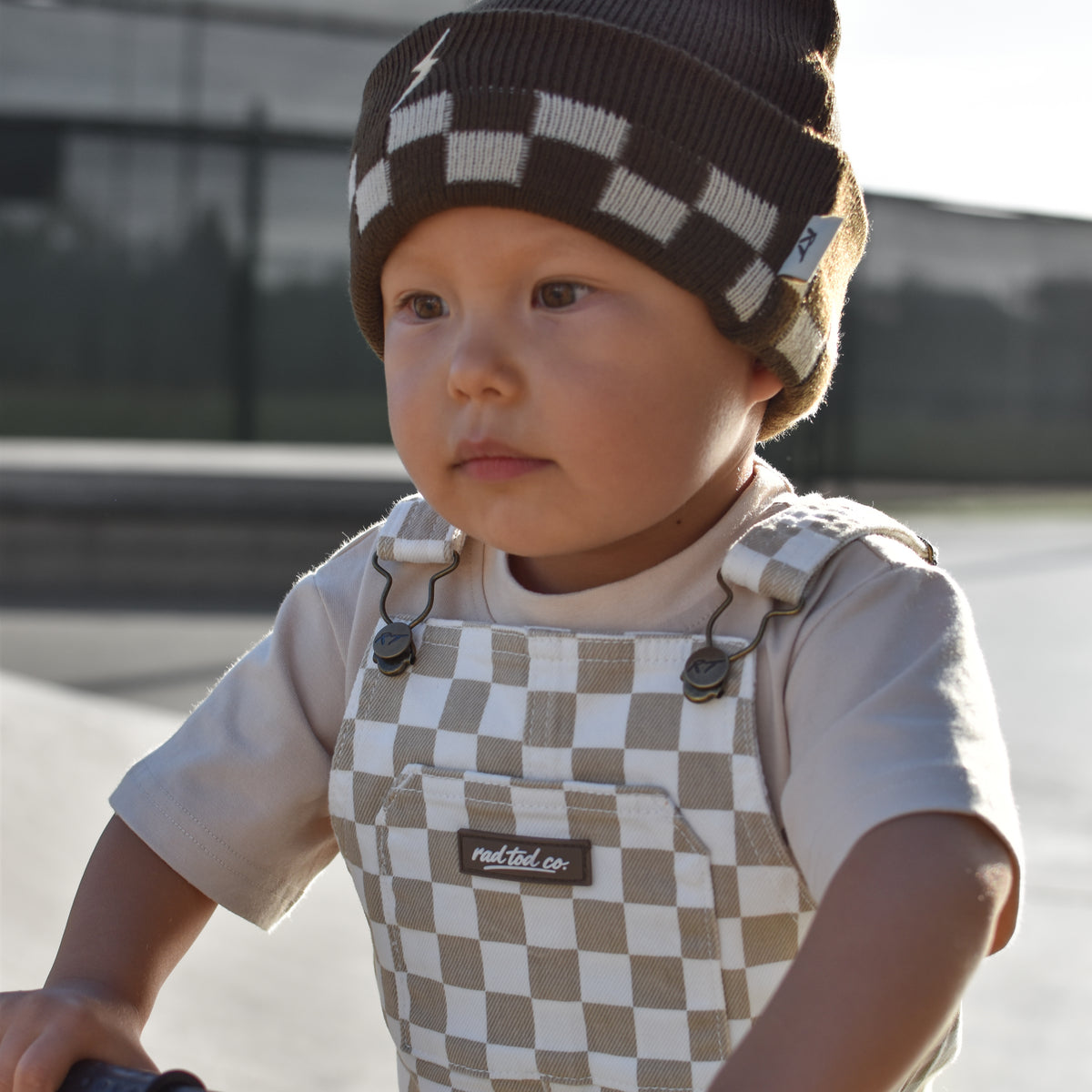 A toddler wearing a brown and beige checkered knit beanie and matching checkered overalls rides a blue bicycle outdoors.