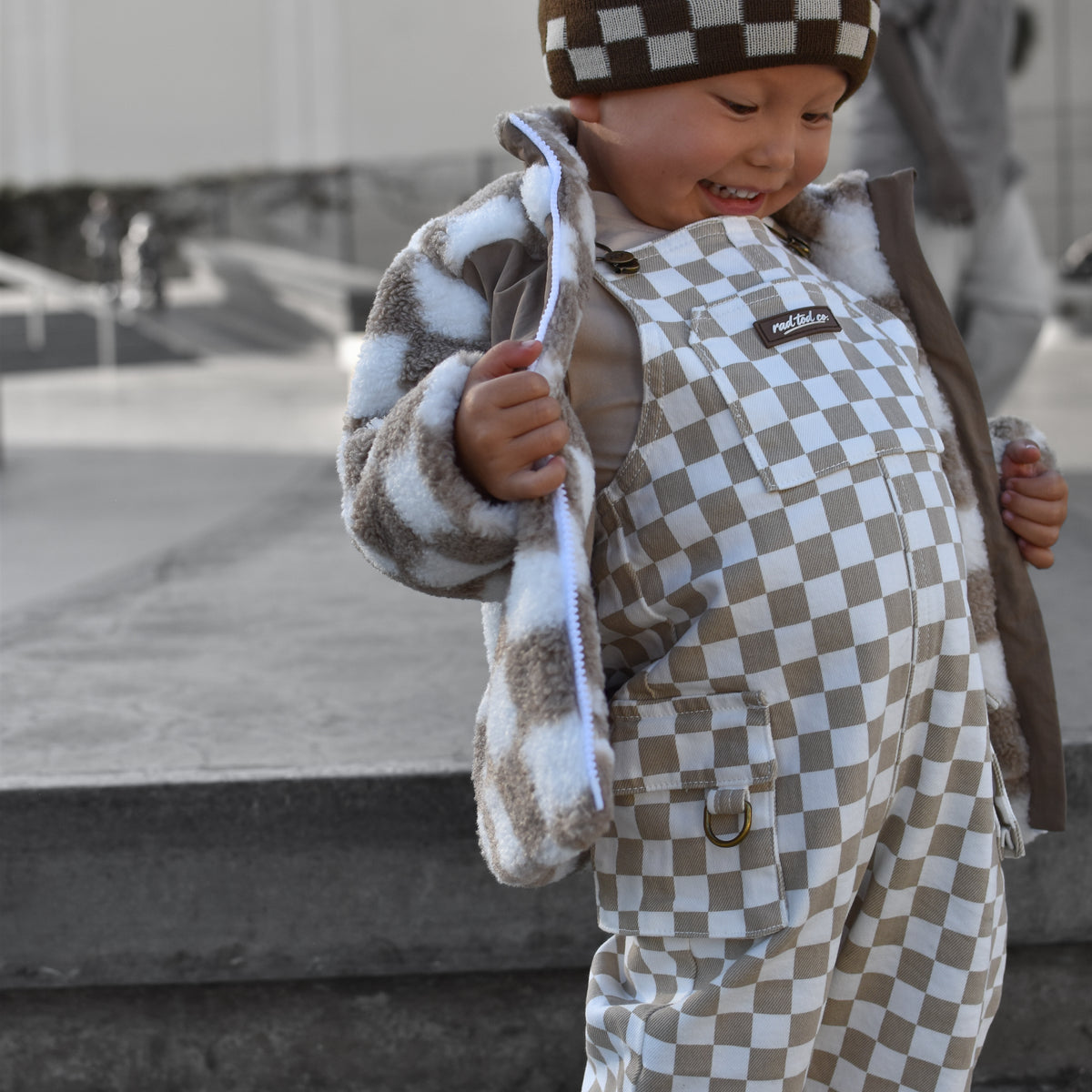 A toddler wearing a brown and beige checkered beanie, a brown and beige checkered overalls, and a brown and white checkered fleece jacket, standing outdoors in a city setting.
