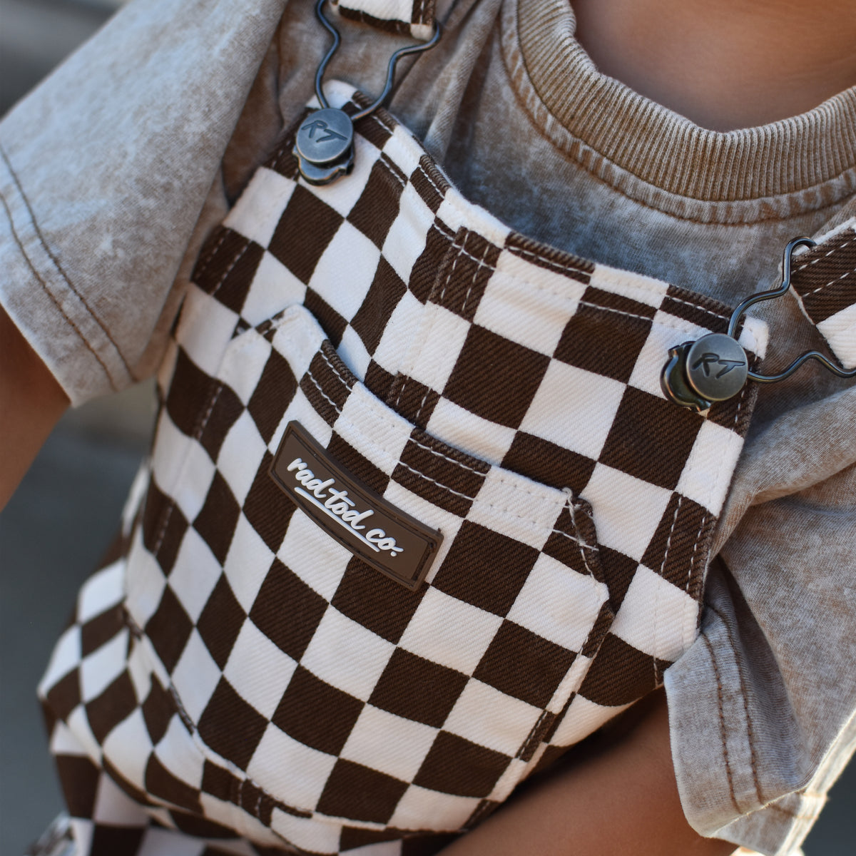 A close-up of a child wearing brown and cream checkered canvas overalls over a faded beige t-shirt. The brand name Rad Tod Co.