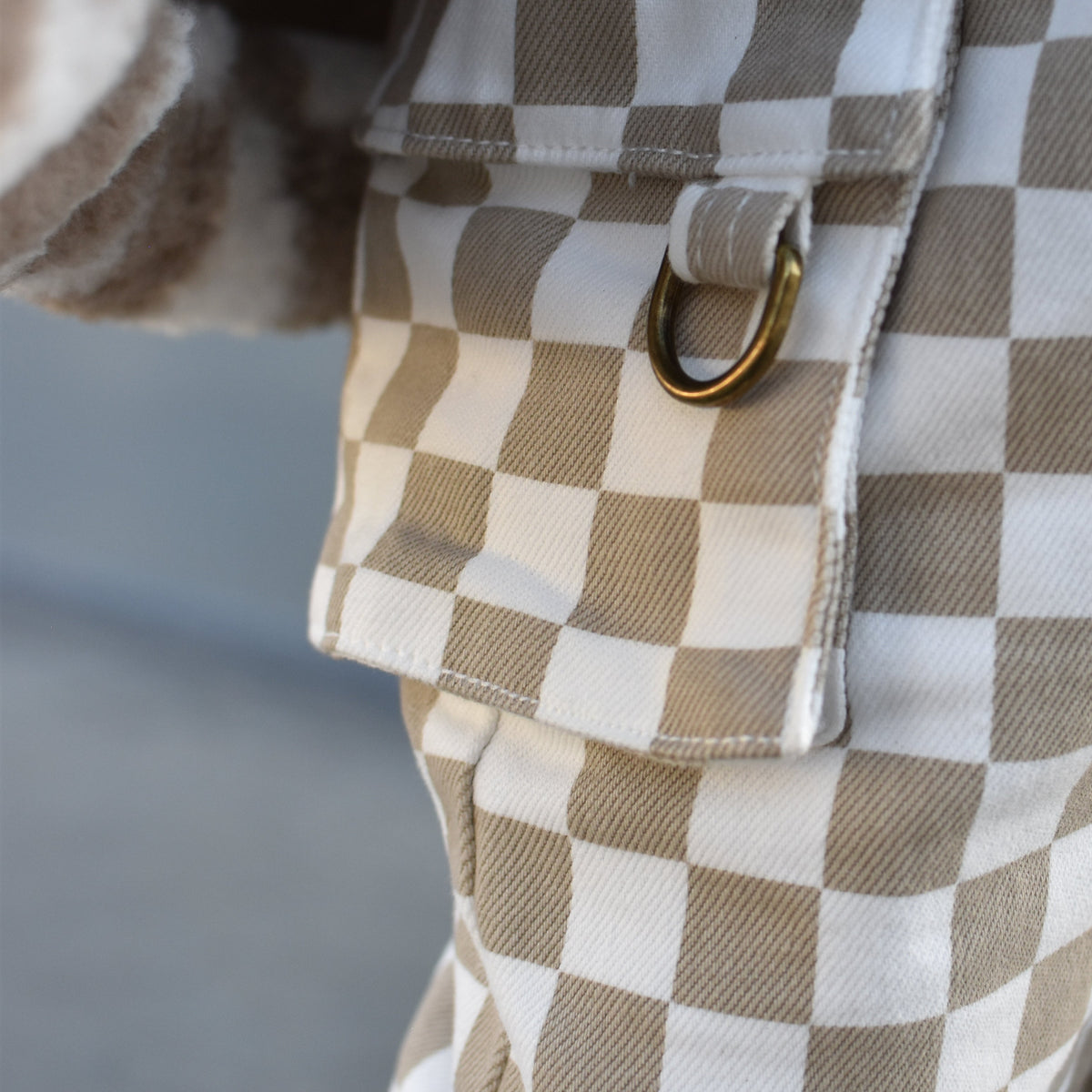 Close-up of a brown and white checkered garment with a flap pocket and a gold-tone metal ring detail.
