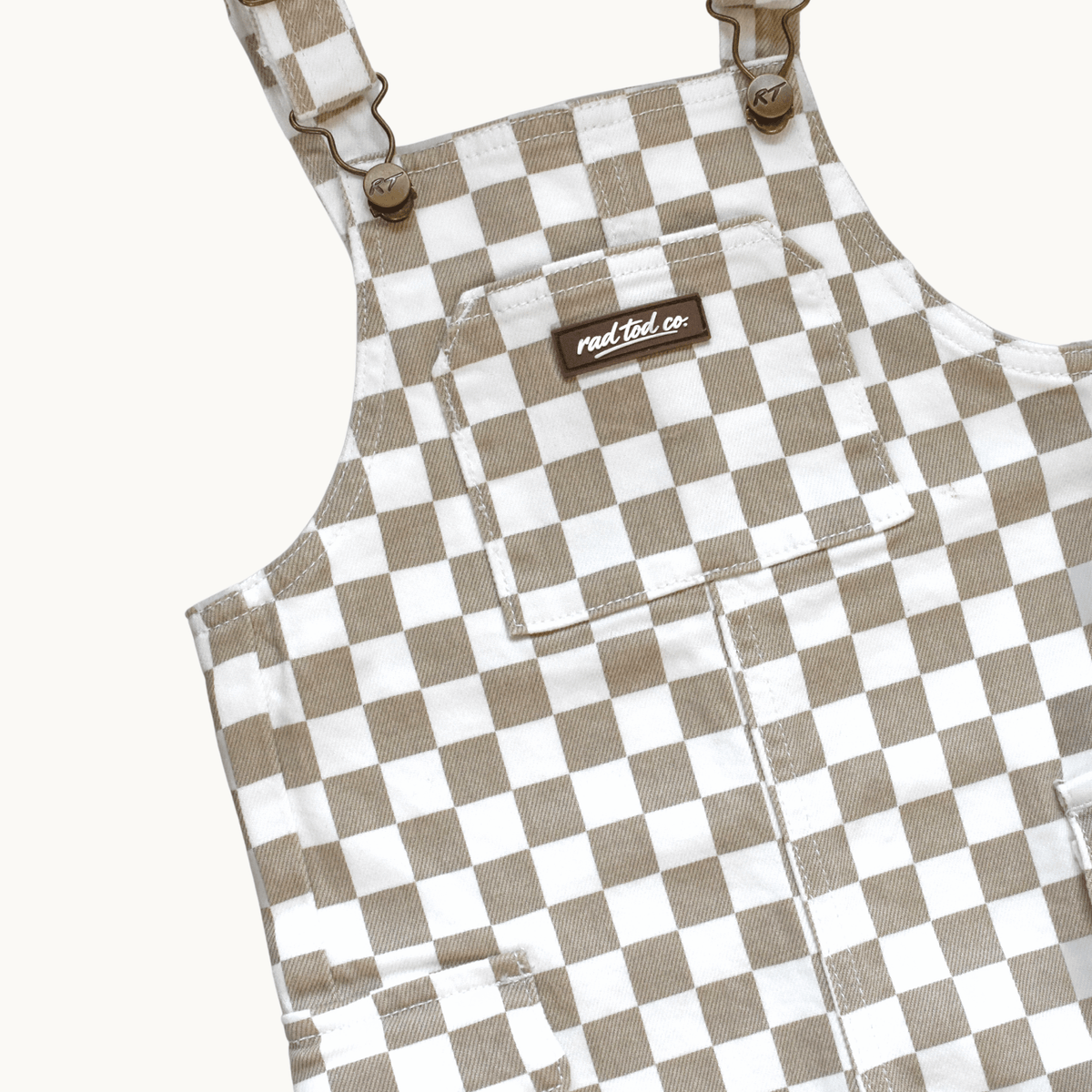 A close-up of a pair of checkered overalls in a beige and white gingham pattern, featuring adjustable shoulder straps with metal buckles, a front chest pocket, and a small brown brand label reading rad tod co,