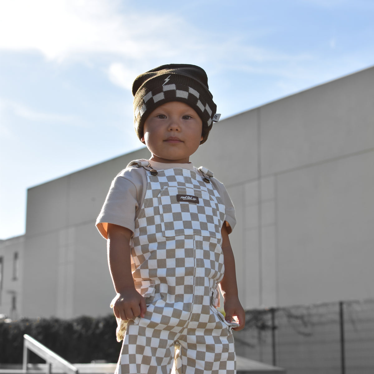 A toddler standing outdoors in front of a modern building, wearing a matching beige and white checkerboard outfit with a small hat.