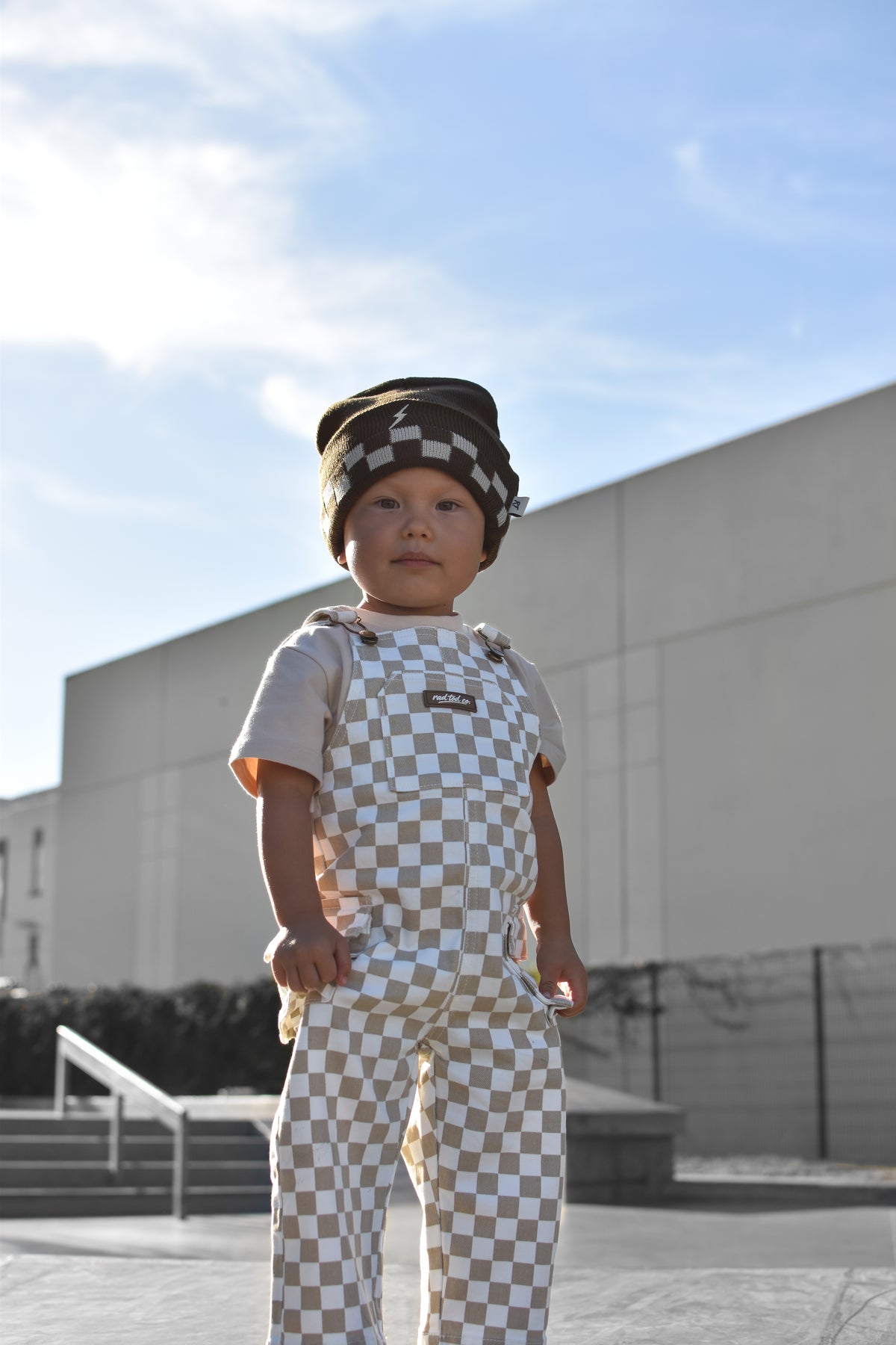 A toddler standing outdoors in front of a modern building, wearing a matching beige and white checkerboard outfit with a small hat.