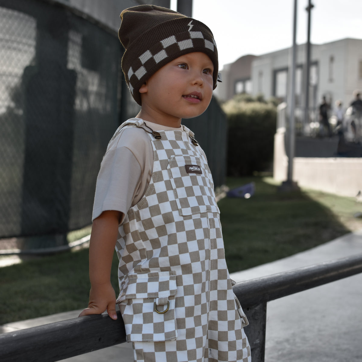 A toddler standing outdoors on a concrete path, wearing brown and beige checkered overalls over a light beige t-shirt and a matching checkered beanie.