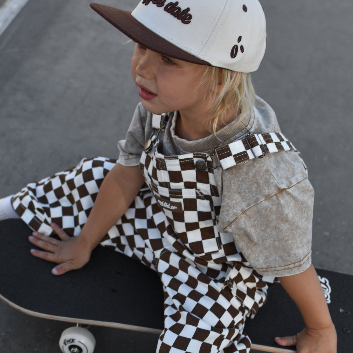 A child wearing brown and white checkered overalls and a matching baseball cap sits on a skateboard on an asphalt surface.
