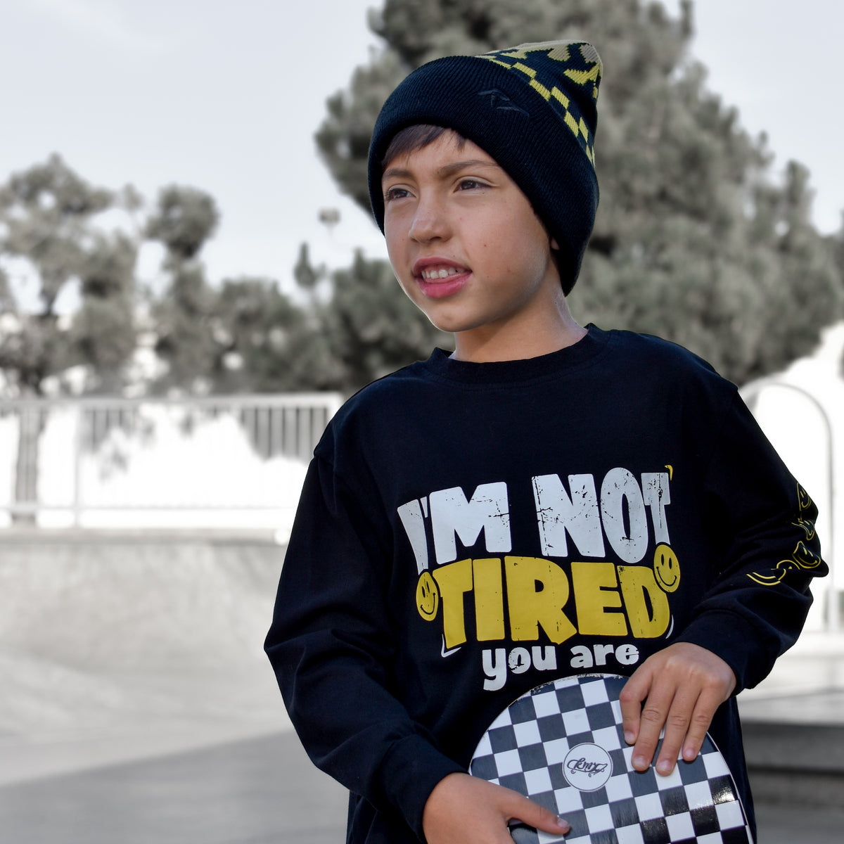 A young boy wearing a black beanie with yellow accents and a black graphic sweatshirt that reads 'I'M NOT TIRED you are' holds a checkered skateboard in an outdoor skatepark setting.